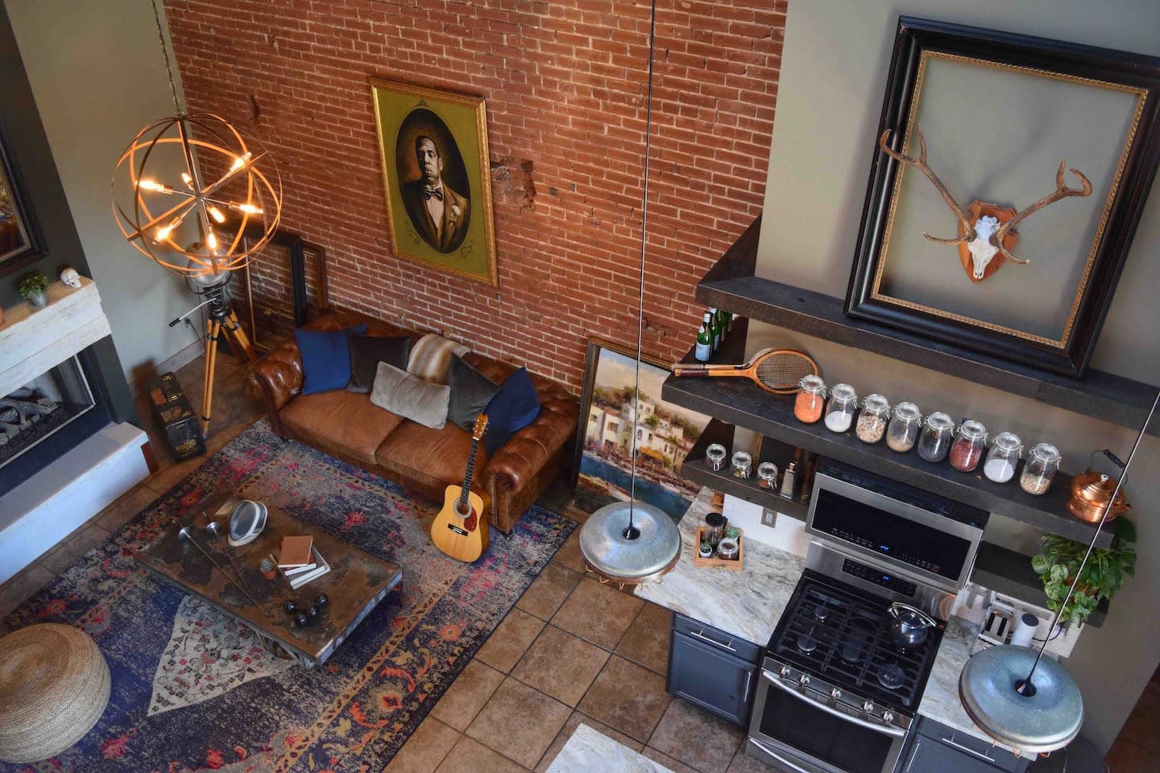 A cozy living space with a leather sofa, an acoustic guitar, a large area rug, and a kitchen area featuring modern appliances and hanging light fixtures, all framed by a brick wall.