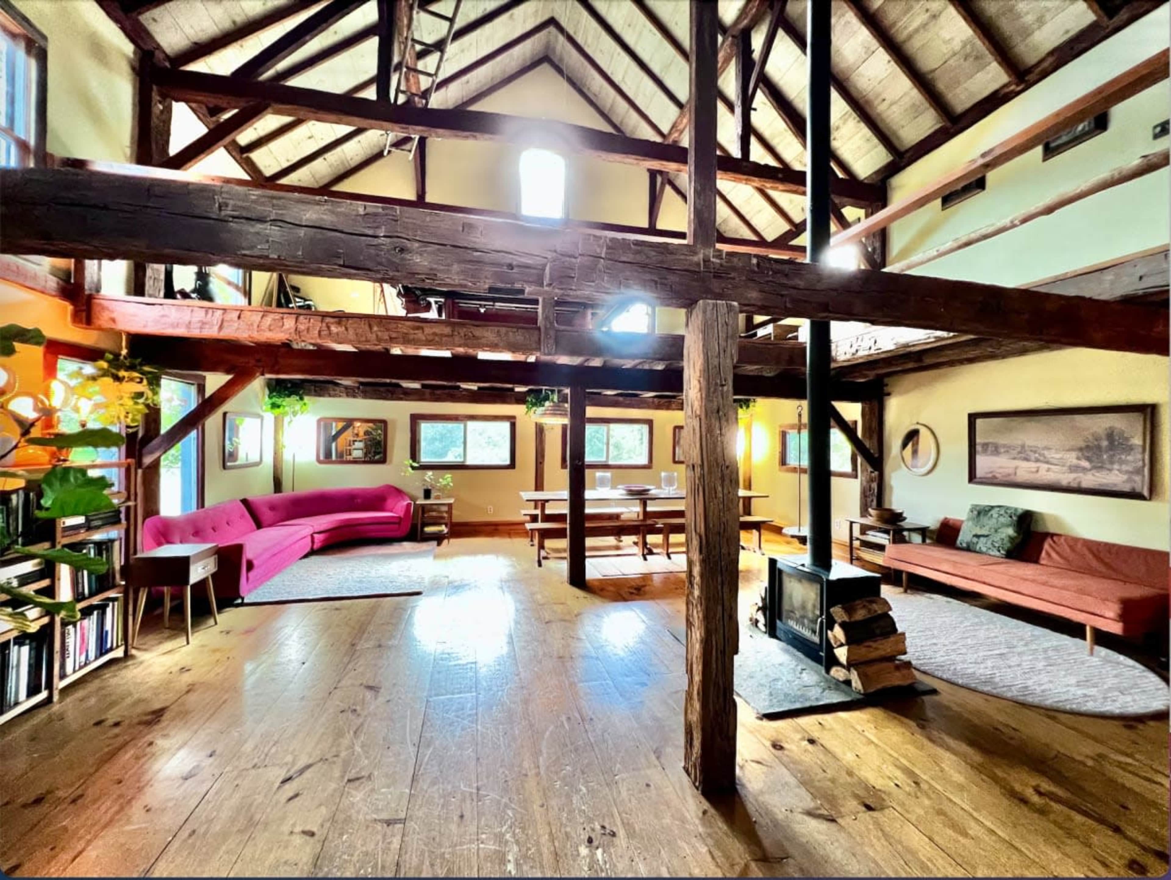 The image depicts a spacious interior of a rustic cabin with high wooden beams, a central fireplace, and a mix of lounge and dining areas featuring a pink couch and wooden tables.