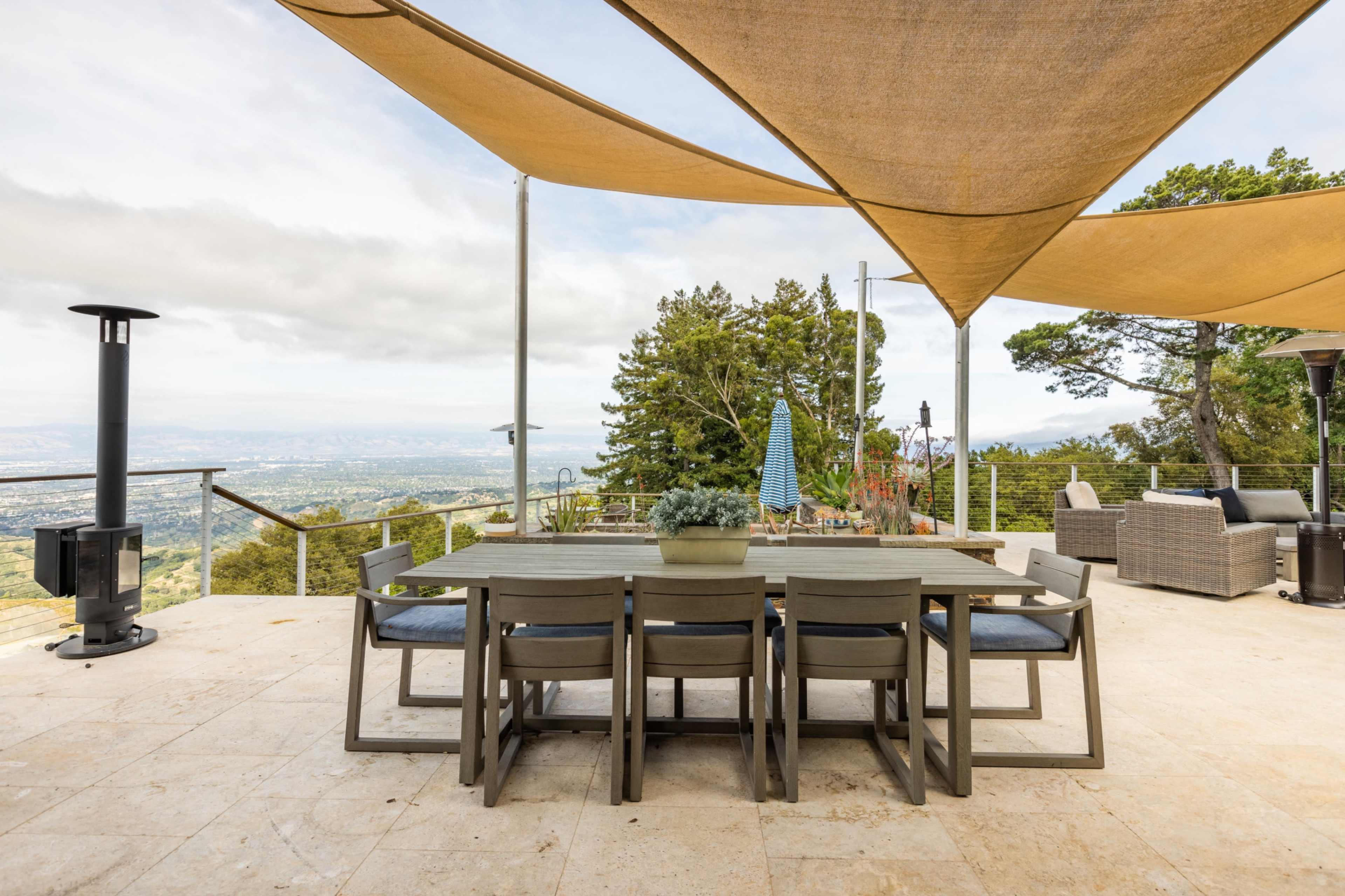 A spacious outdoor dining area features a large table surrounded by chairs, shaded by fabric canopies, with a view of a valley and distant mountains.