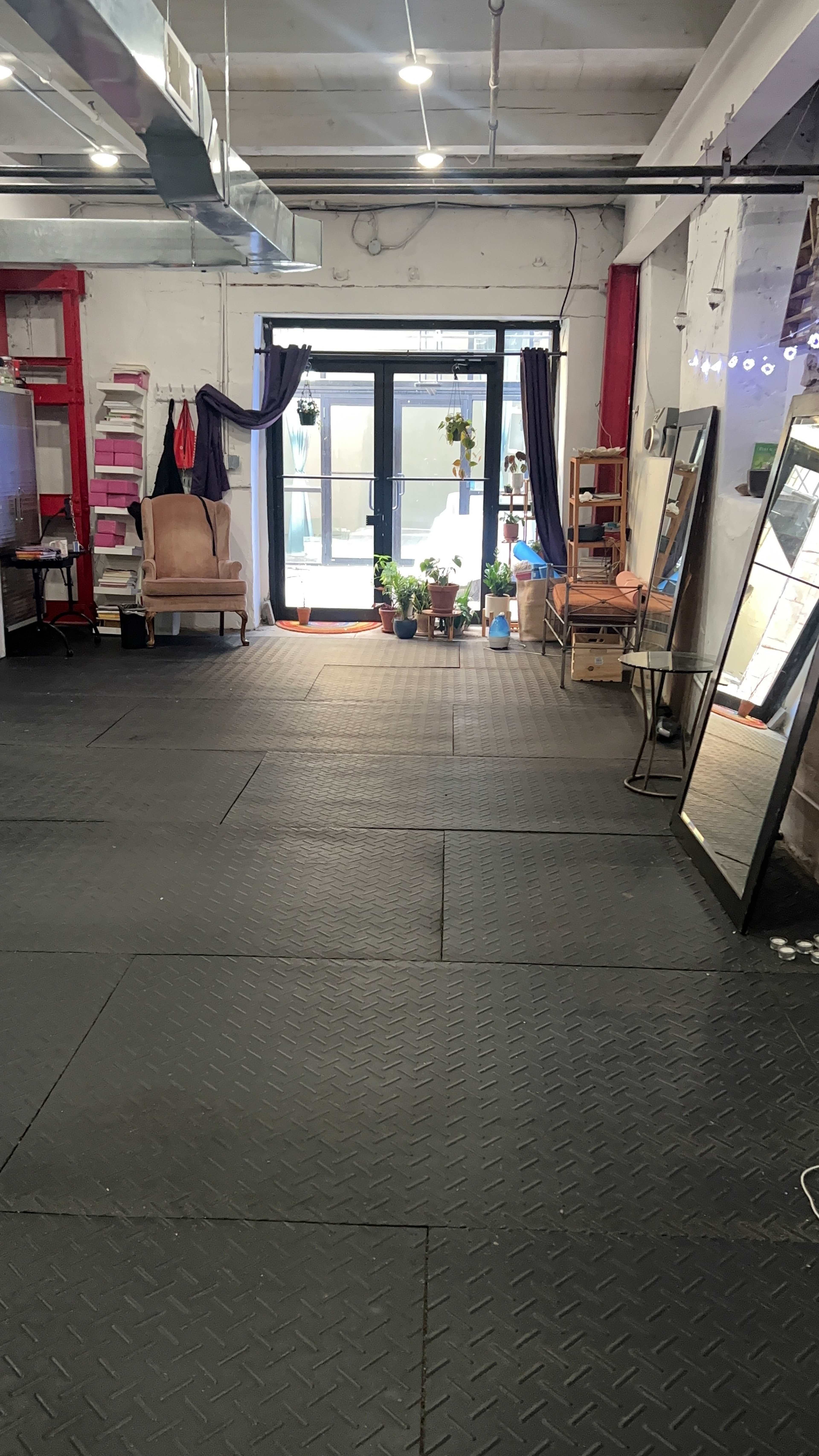 An open interior space with black rubber flooring, a chair, mirrors, and several potted plants near a large glass door.