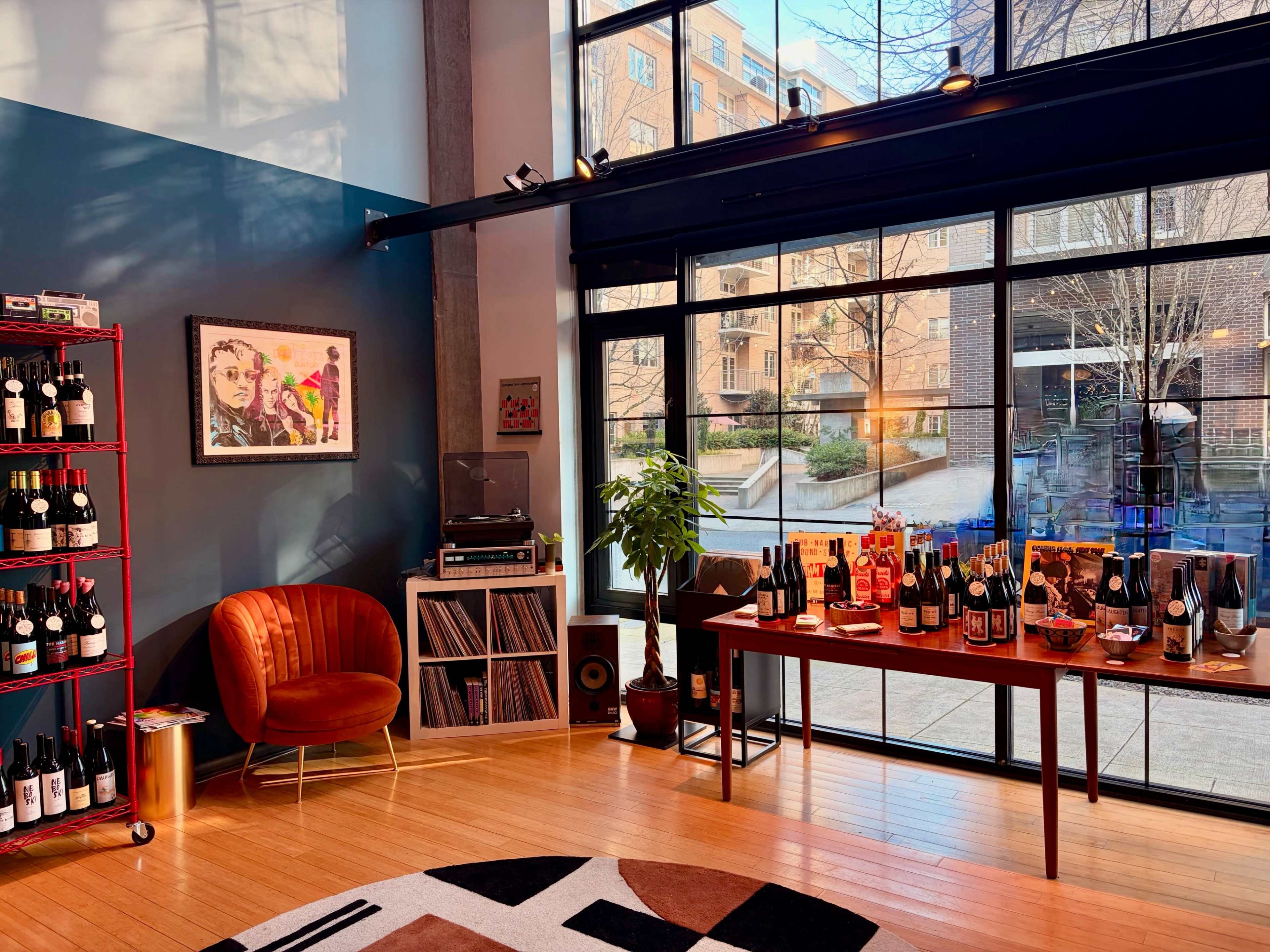The image shows a modern interior space featuring a display of wine bottles on shelves, a vinyl record setup, and a large window revealing an outdoor view.