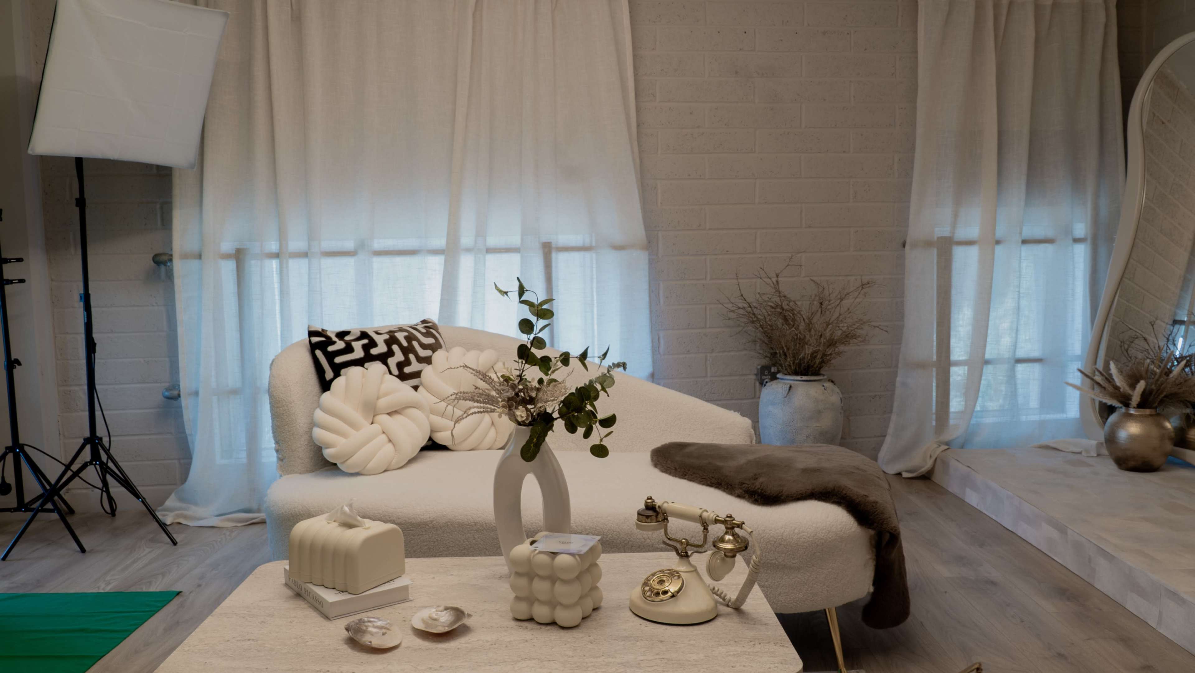 The image shows a modern, minimalist living room with a white curved couch, decorative pillows, a coffee table featuring various items, and sheer curtains filtering natural light.