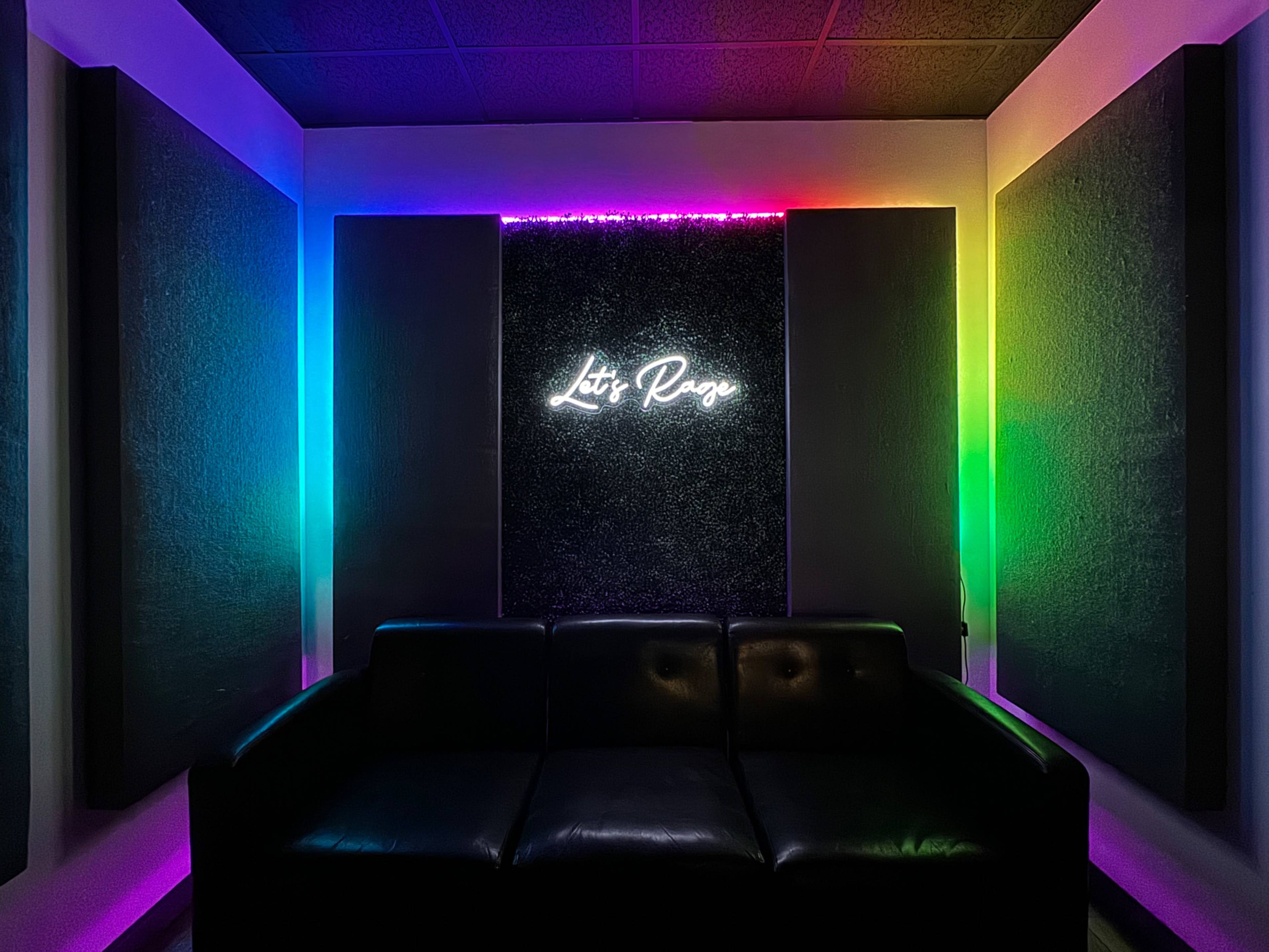 A dimly lit room featuring a black leather couch and a neon sign that reads "Lit Page," with colorful LED lighting on the walls.