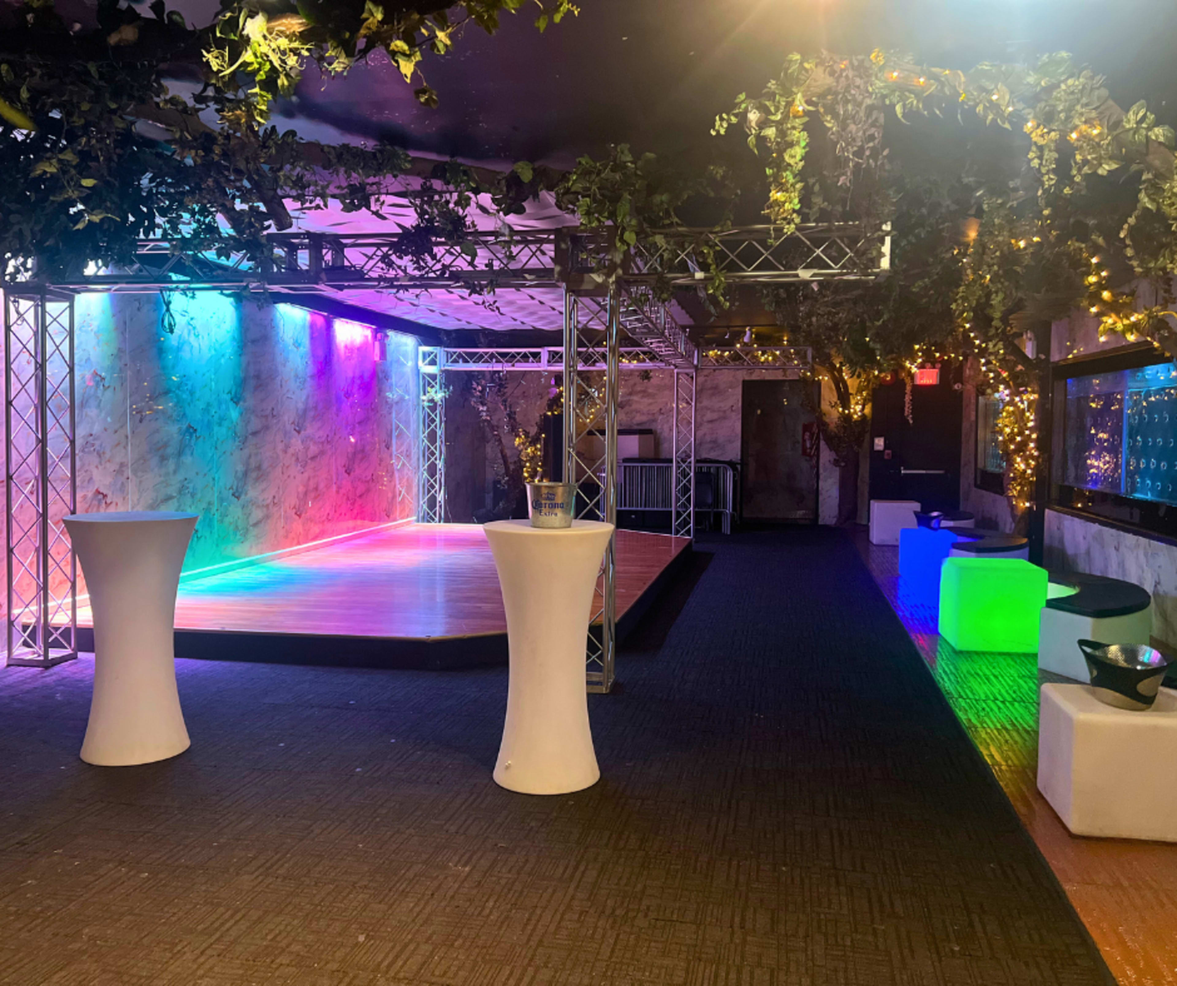 The image shows a vibrant event space with a small stage illuminated by colorful lights and decorated with greenery, surrounded by white and colorful seating options.
