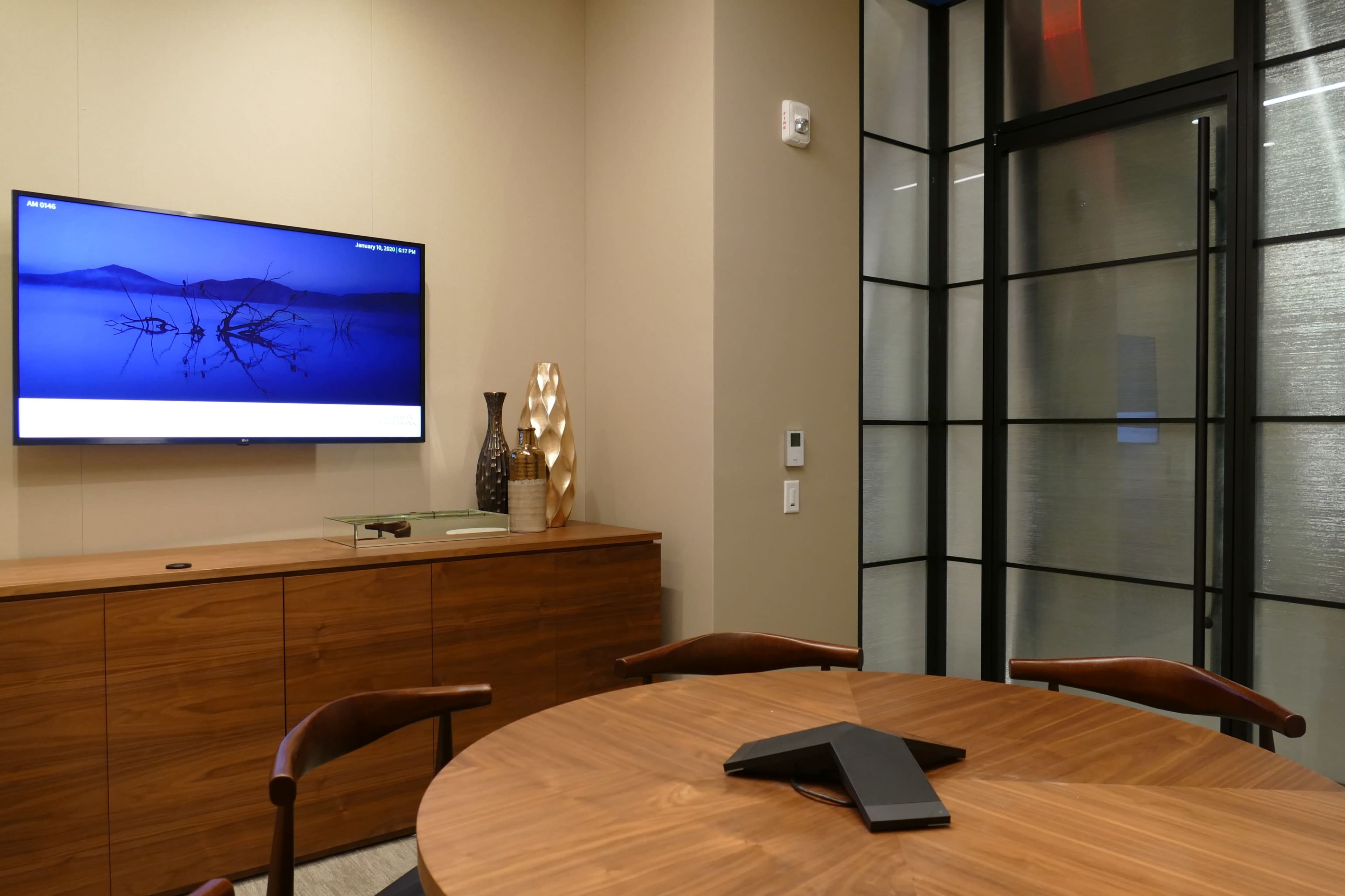 A conference room features a round wooden table, a wall-mounted TV displaying a blue image, and a glass door leading to an adjacent space.