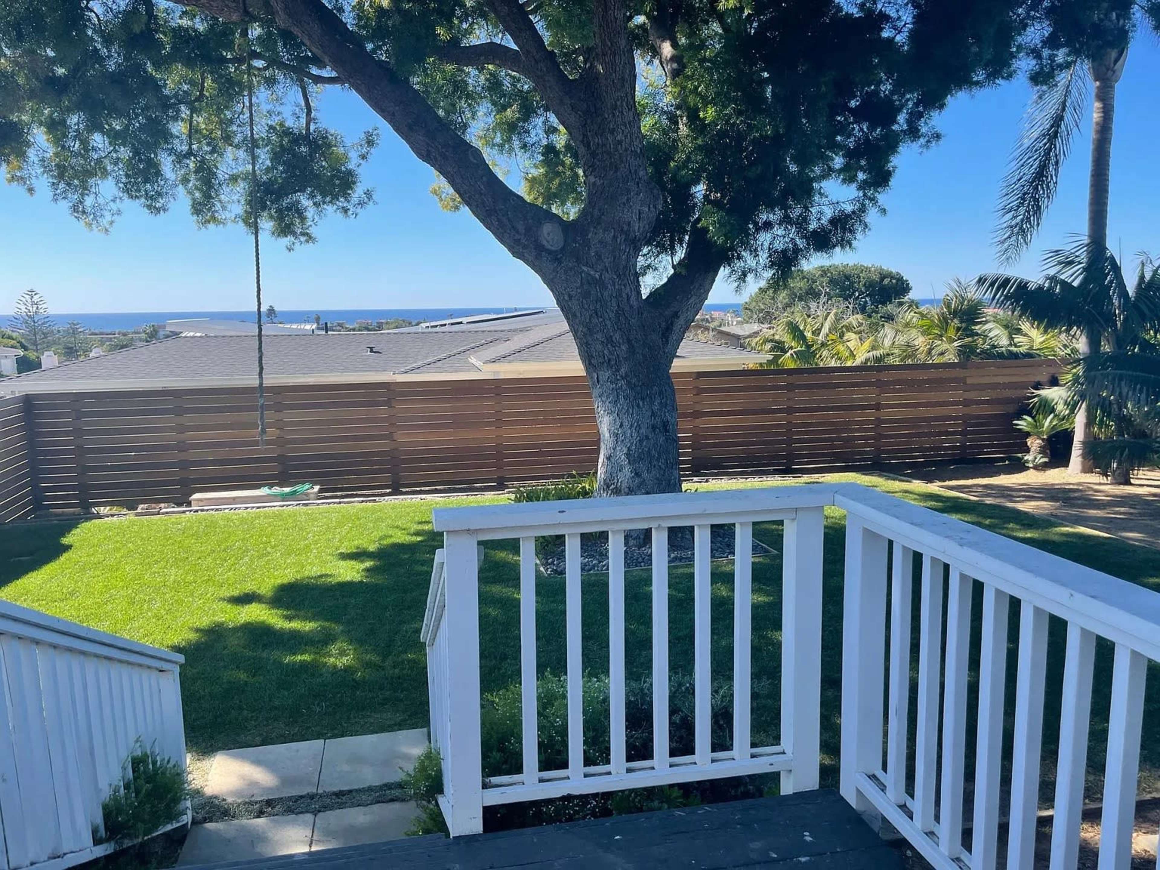 A backyard view shows a lush green lawn, a large tree, and a fence bordering the area, with the ocean visible in the distance.