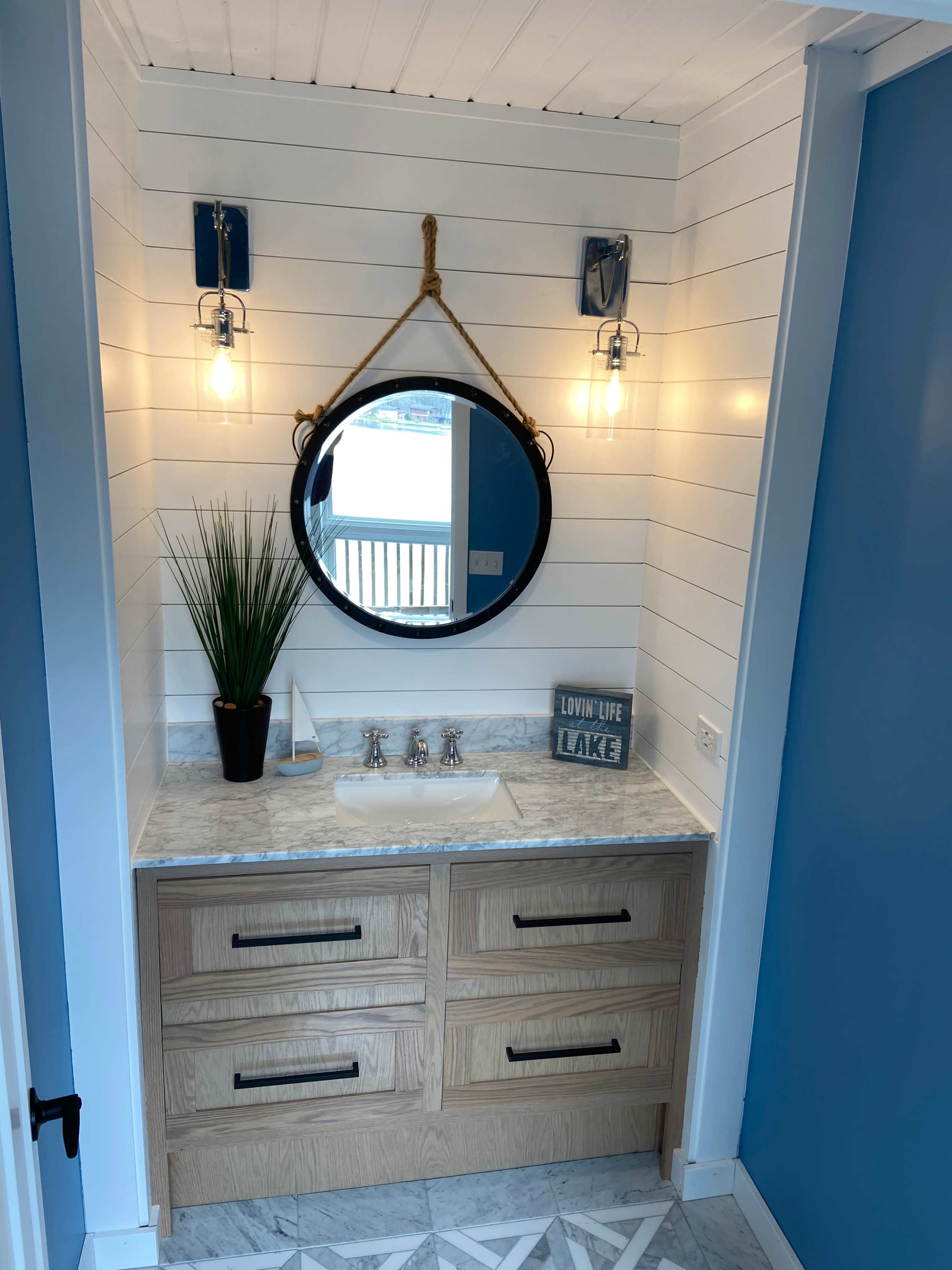A modern bathroom vanity with a round mirror, two wall sconces, a plant, and a stone countertop above wooden drawers, set against a light wood paneled wall and blue accent wall.