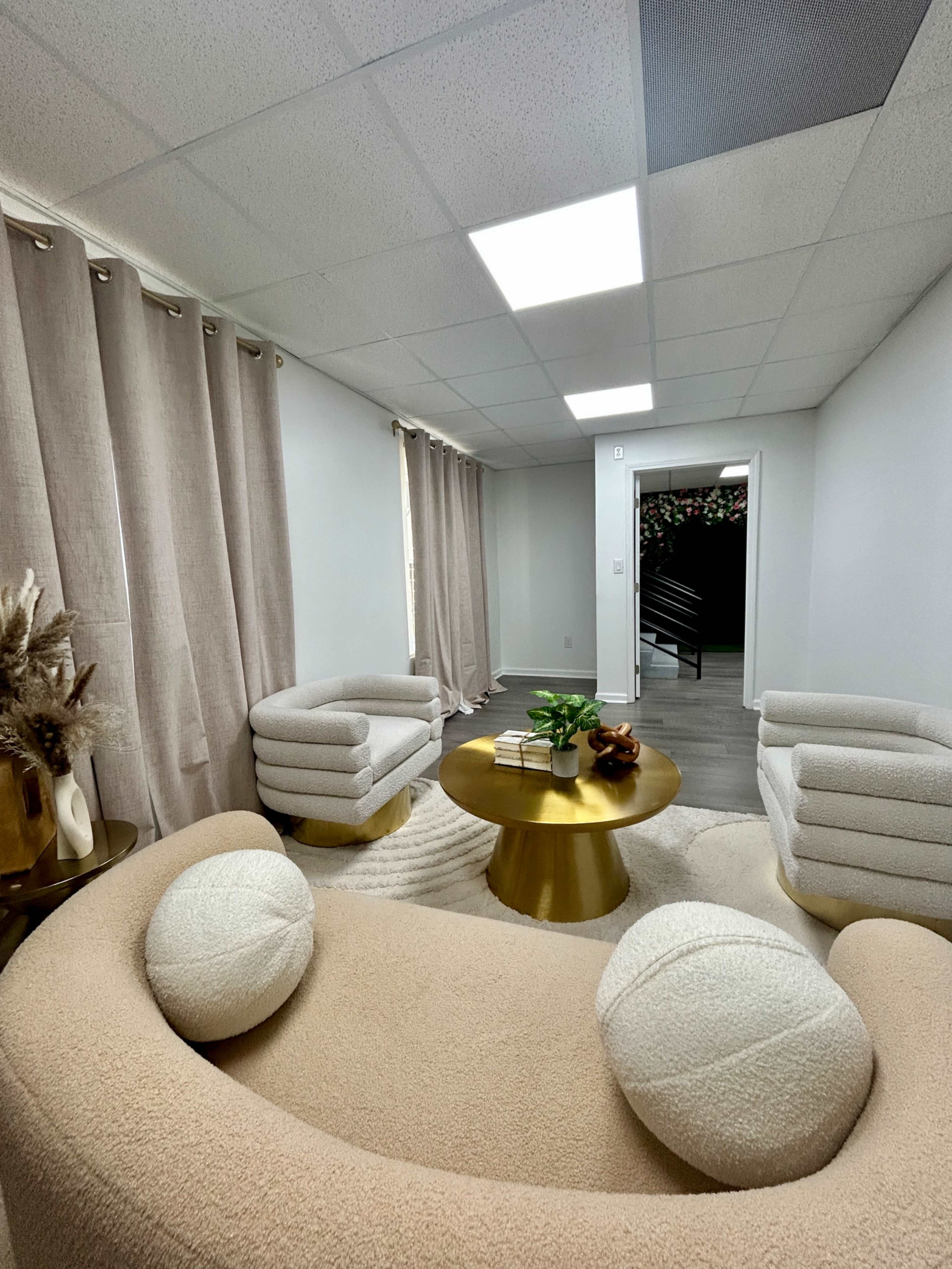 The image shows a modern waiting room with a round gold coffee table, two light-colored upholstered chairs, and a curved sofa, all set against pale walls and light curtains.