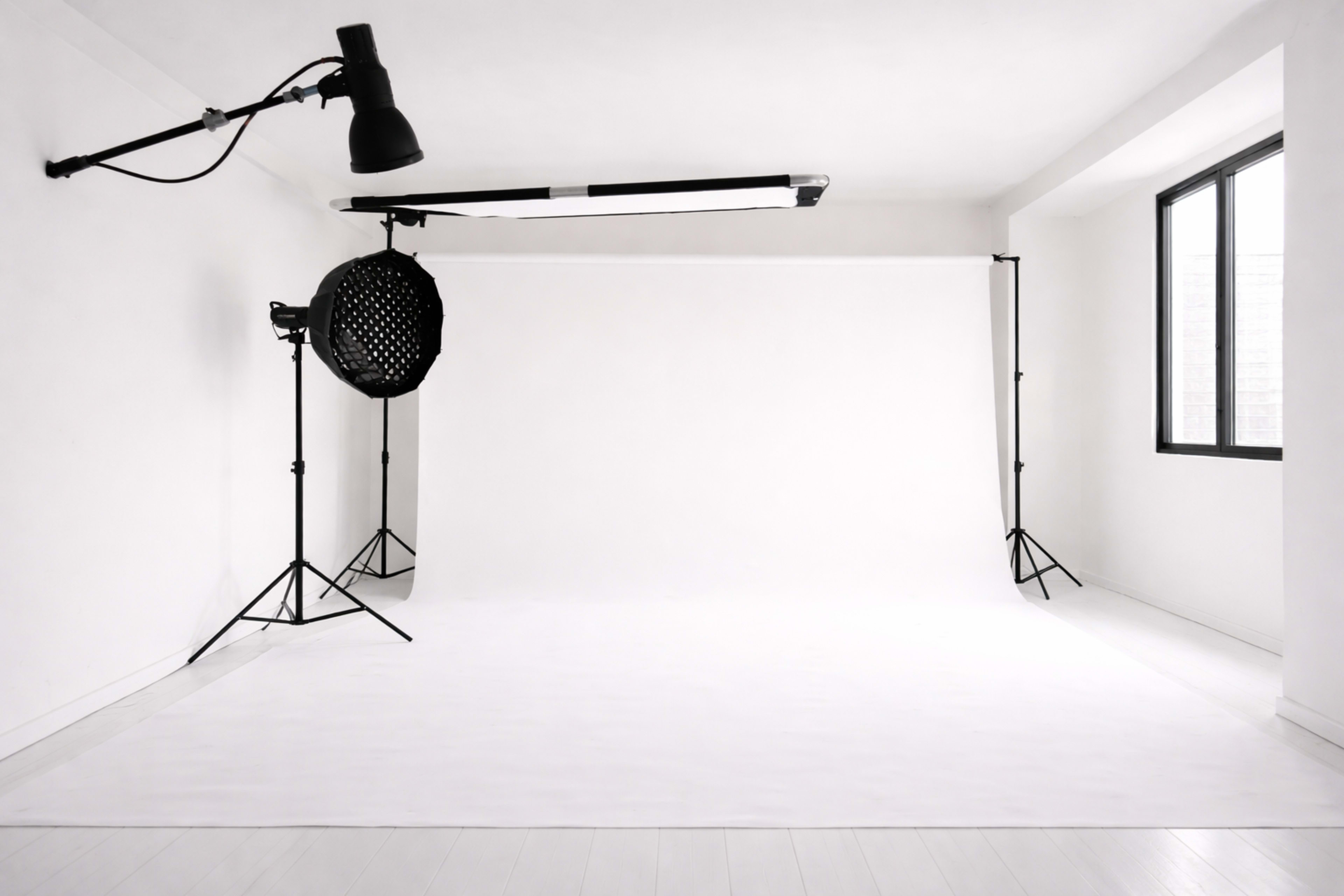 The image shows an empty photography studio with a white backdrop, lighting equipment, and large windows.