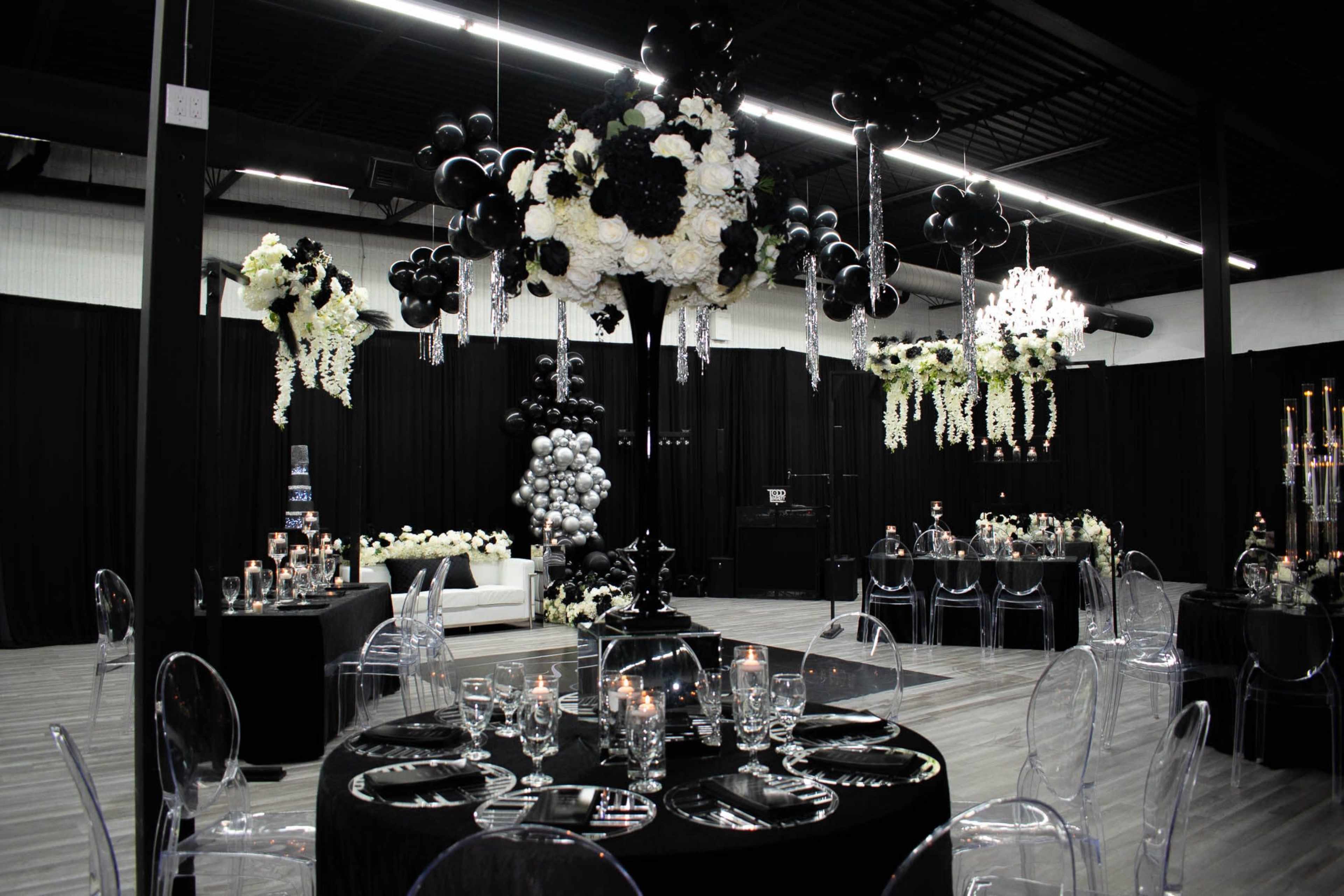 The image shows an elegantly decorated event space featuring black and white floral arrangements, clear acrylic furniture, and tables set with sparkling glassware and candles.