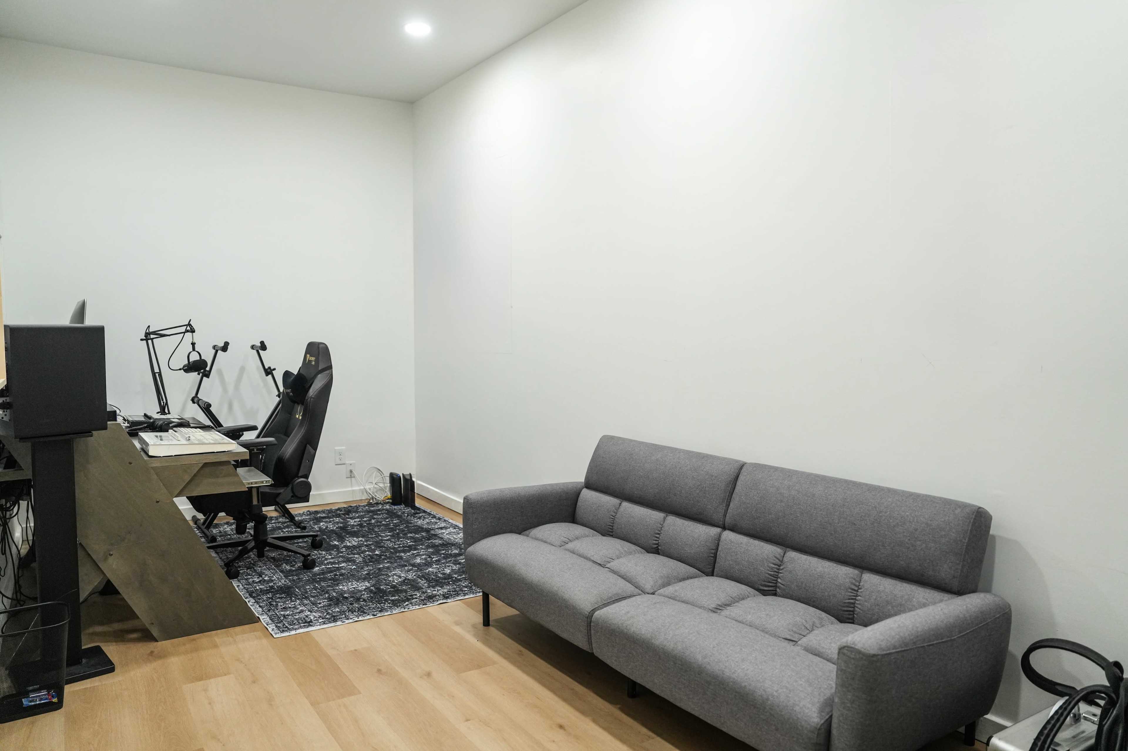 The image shows a minimalist office space featuring a gray sofa, a desk with a computer monitor, and an empty wall with a hardwood floor.