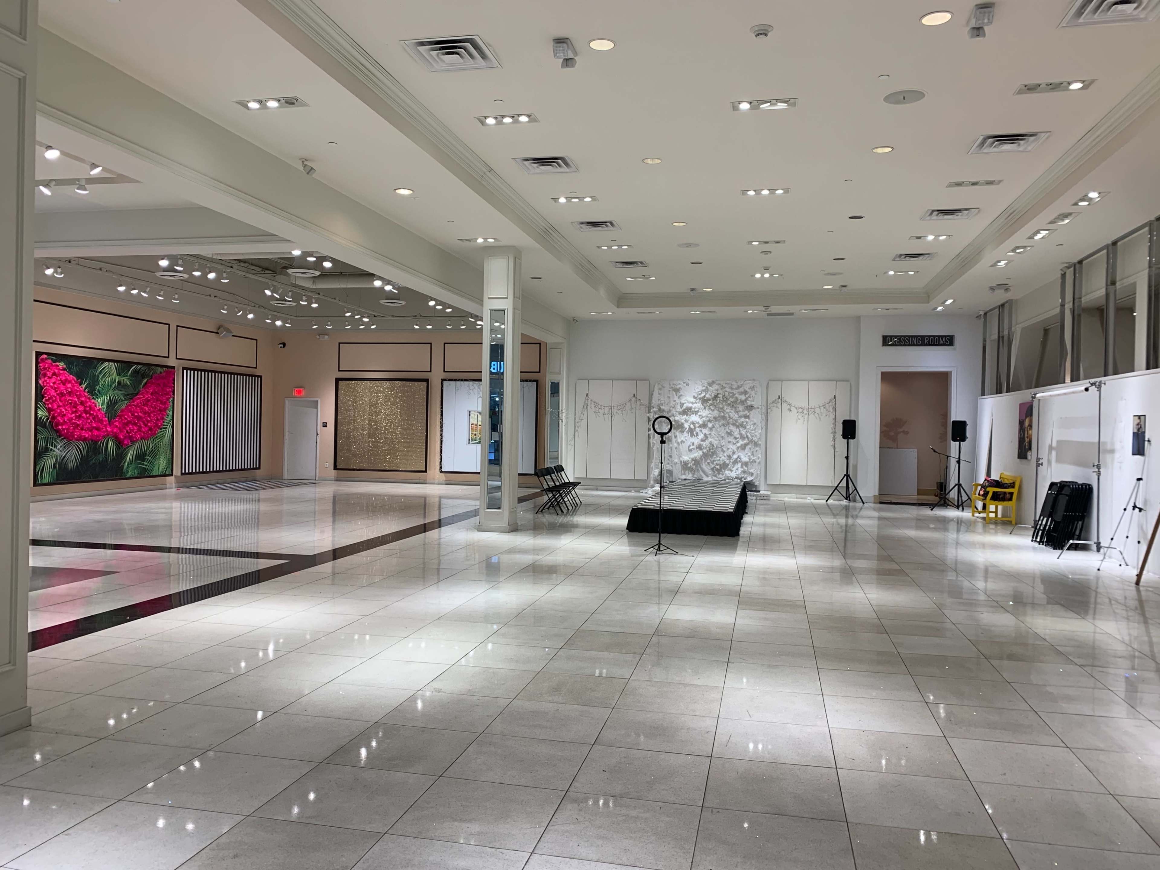 A spacious, well-lit exhibition hall featuring several empty chairs and art displays, with a large floral artwork on one wall.