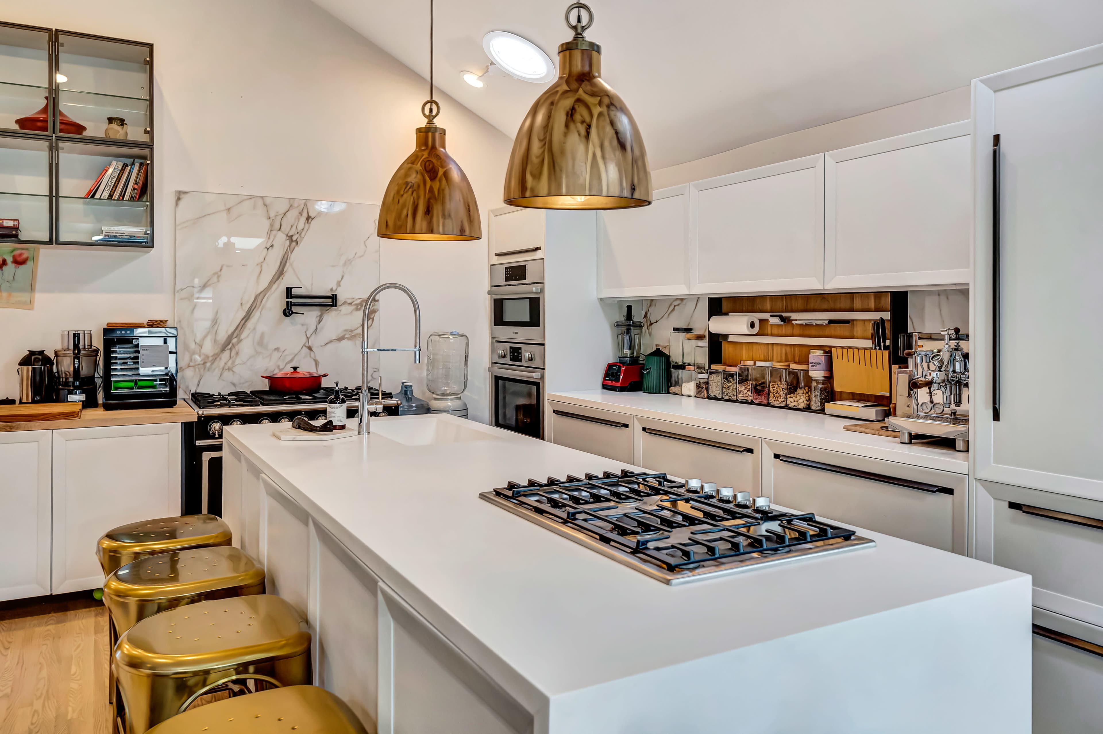 The image shows a modern kitchen featuring a central island with a gas cooktop, two pendant lights above, and white cabinetry lined against the walls.