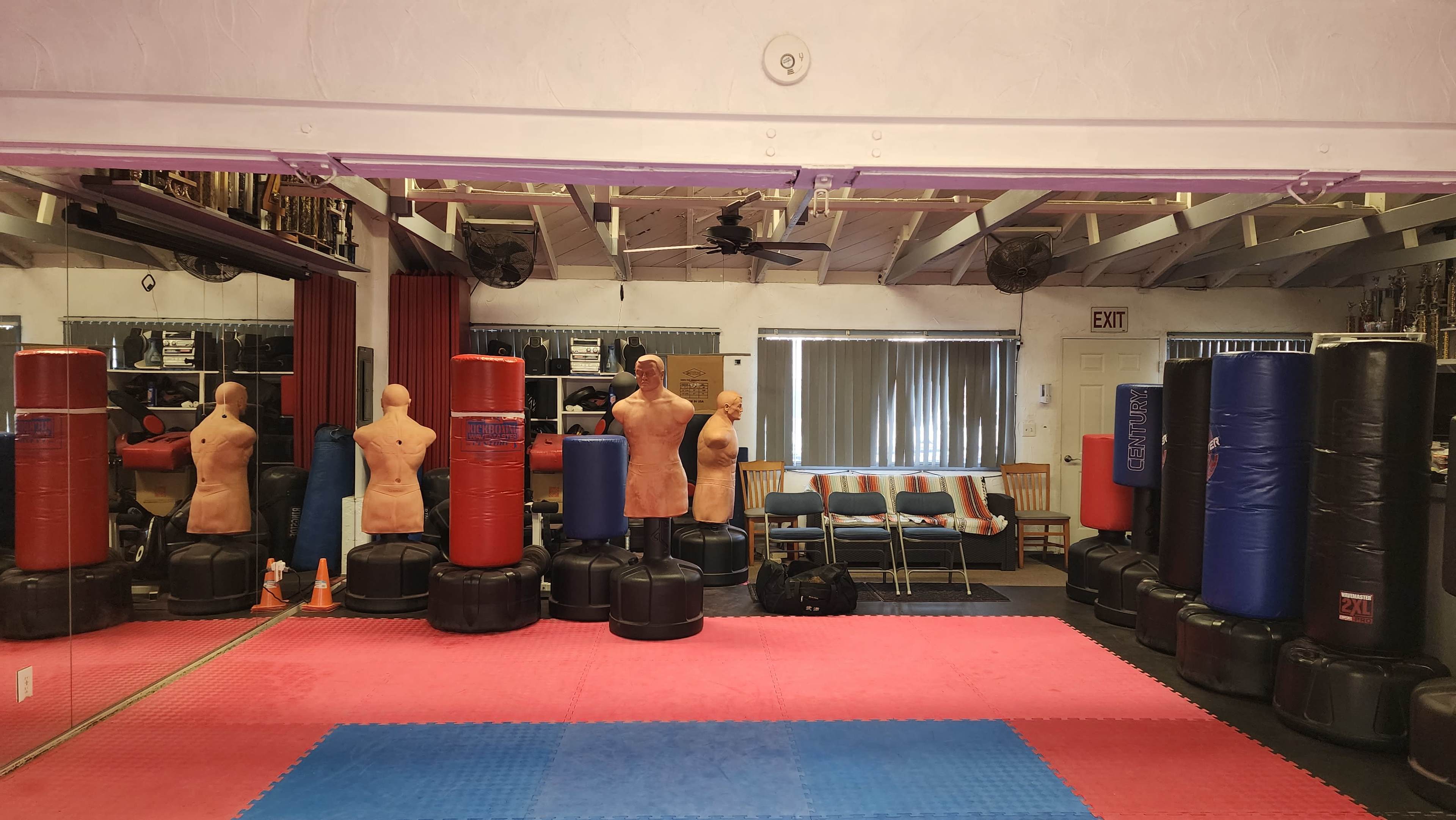 The image shows a martial arts training room with various training dummies and heavy bags arranged in a space featuring red and blue mats.
