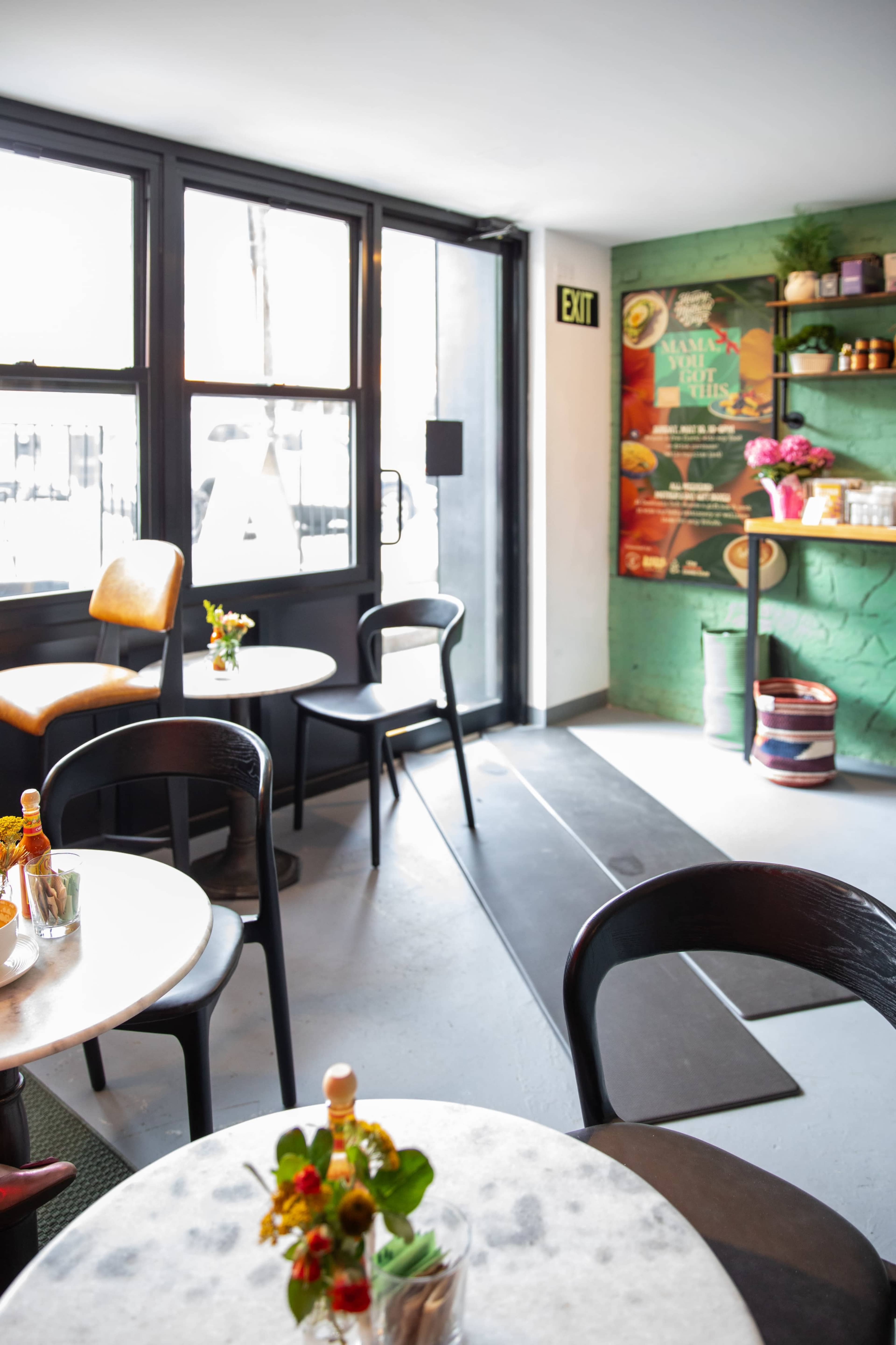 The image shows a cozy café interior with small tables, black chairs, and a decorative green wall featuring promotional artwork.