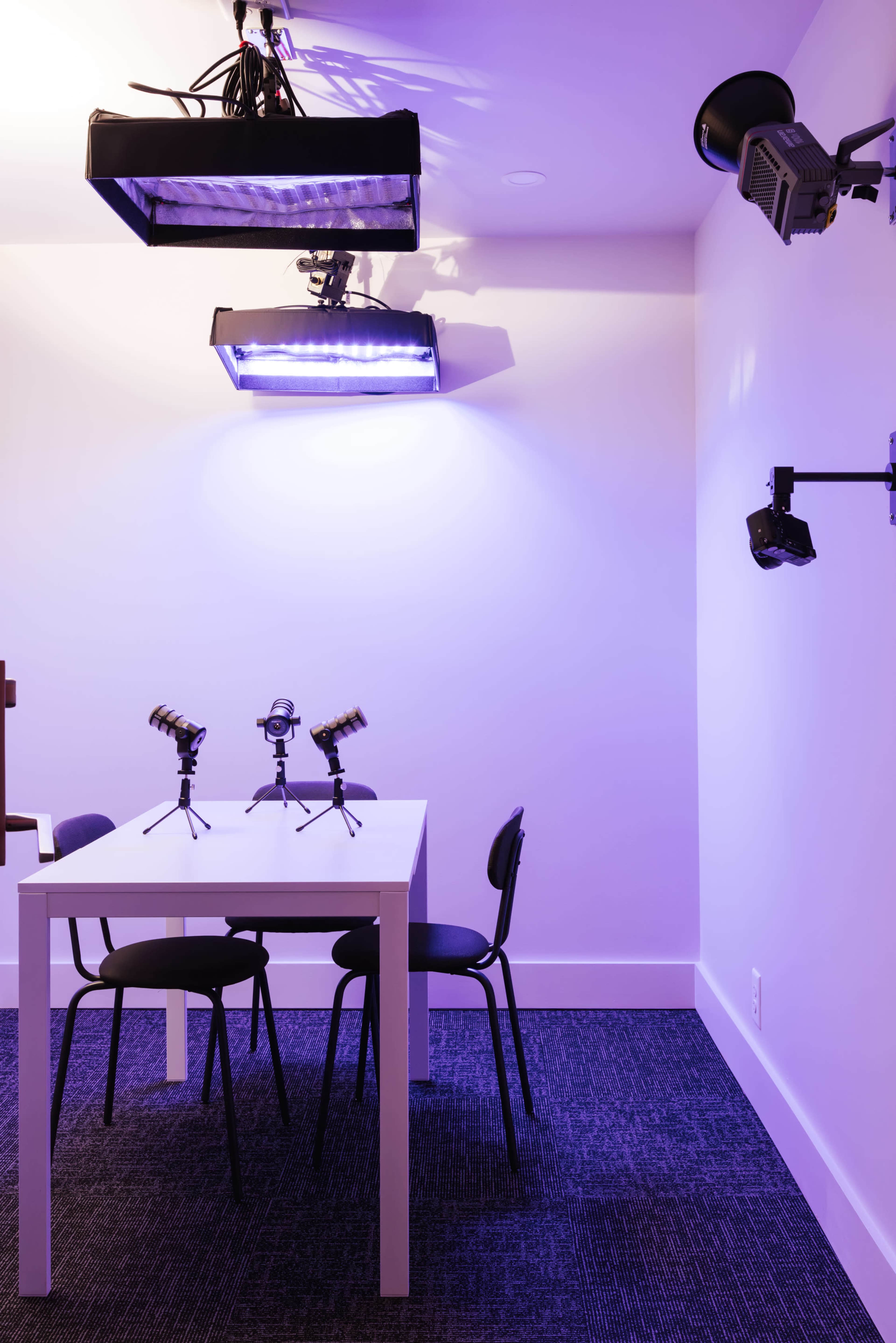 A small, minimally furnished room features a white table with two microphones on stands and several mounted lights on the walls.