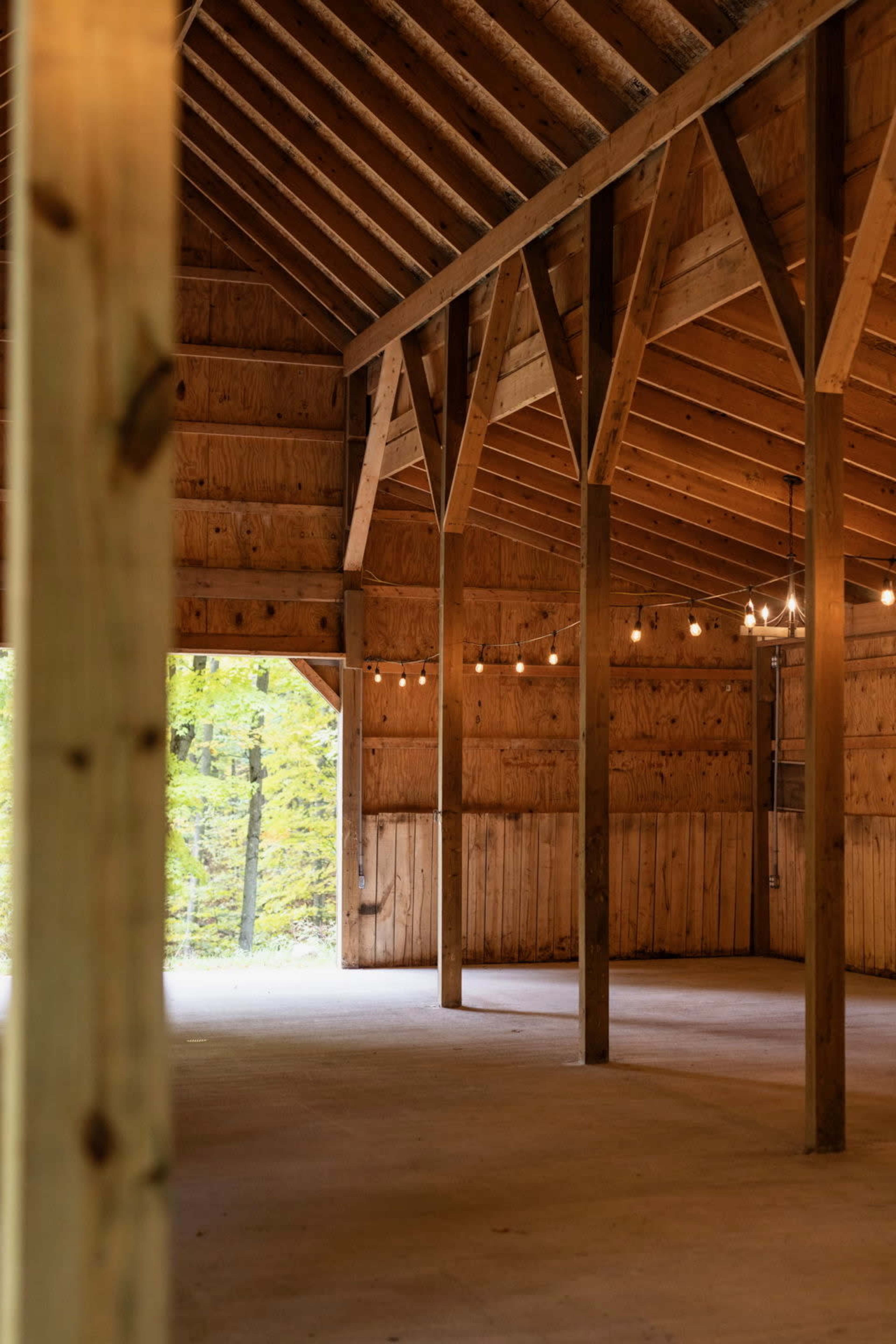 The image shows the interior of a wooden structure with exposed beams and string lights, featuring bare walls and a concrete floor, while a view of trees is visible through an open doorway.