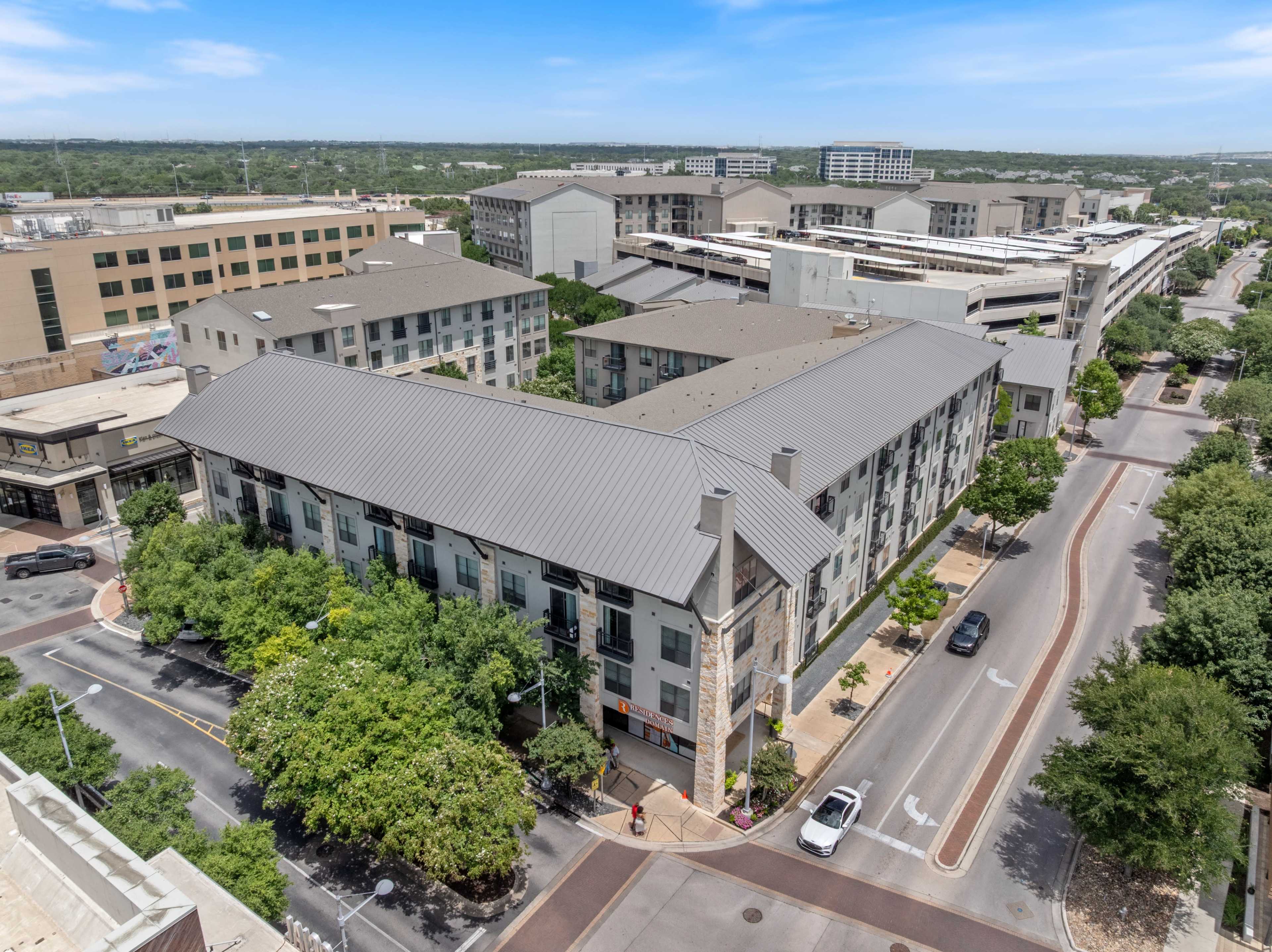 The image shows a modern apartment complex surrounded by trees and commercial buildings in an urban area.
