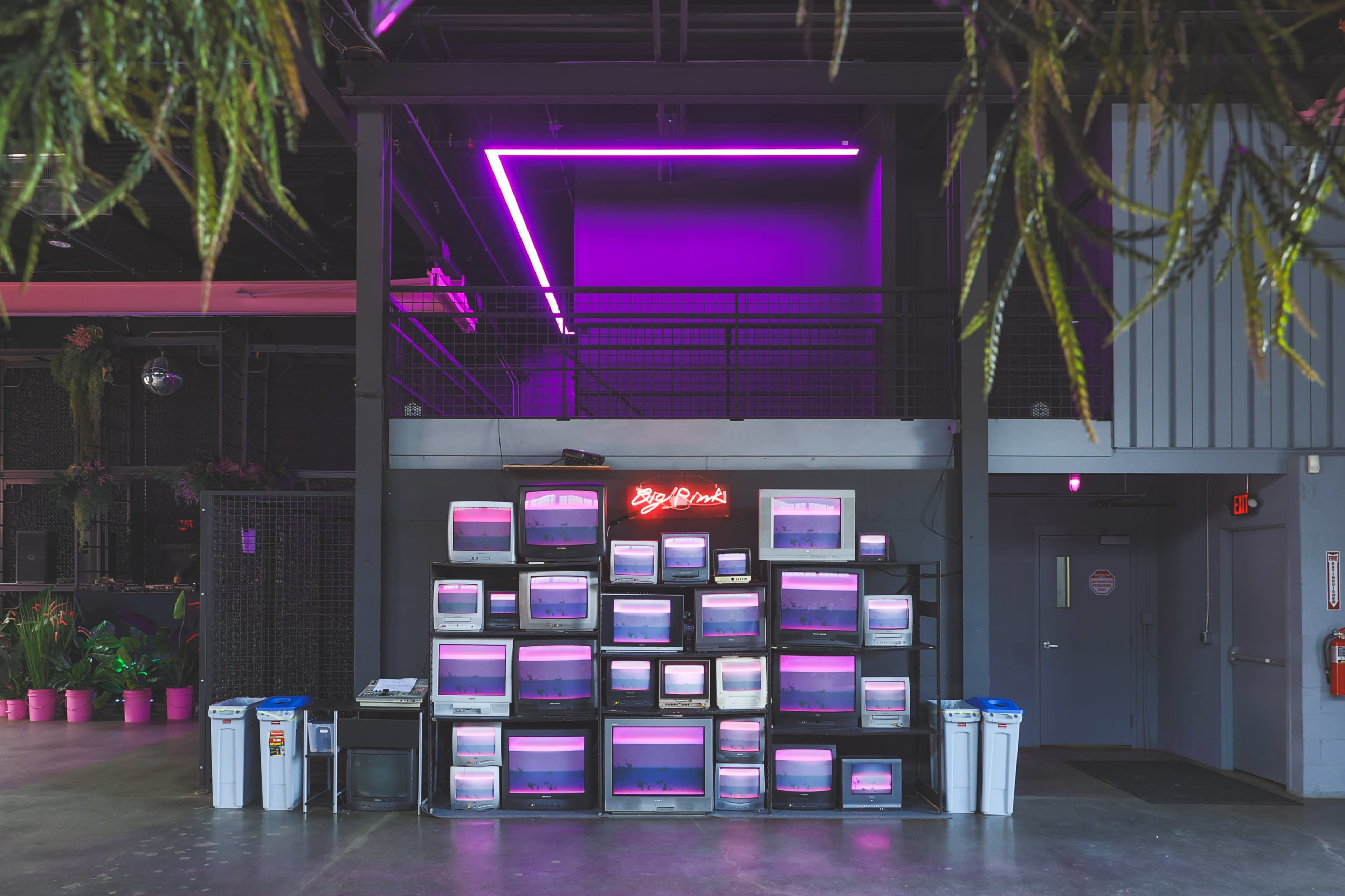A wall of retro computer monitors is arranged in a grid, illuminated by pink neon lights in a modern indoor space.