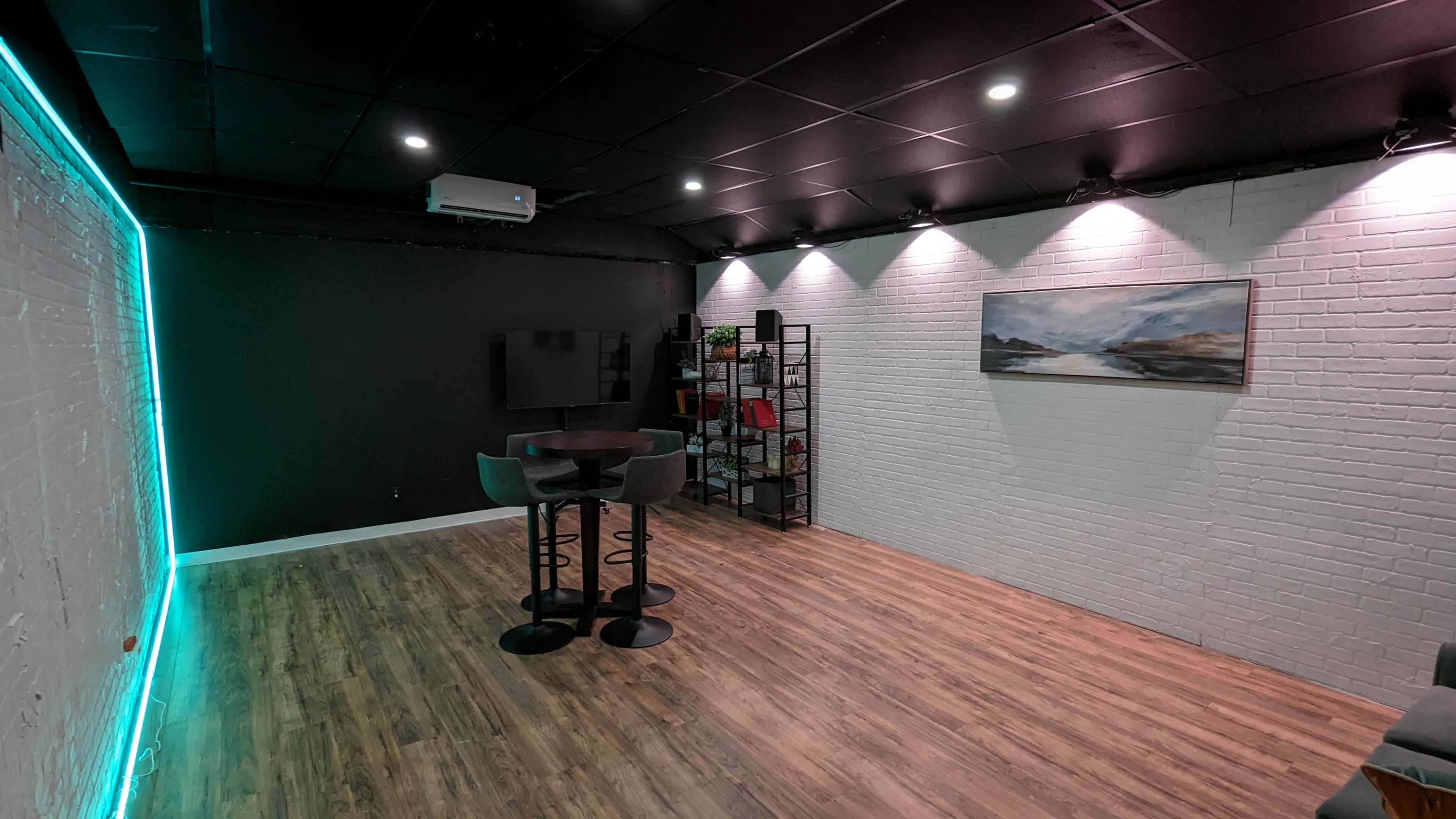 A minimalist room with wooden flooring, a small table and stools in the center, and a black wall with a projector and a shelf against it.
