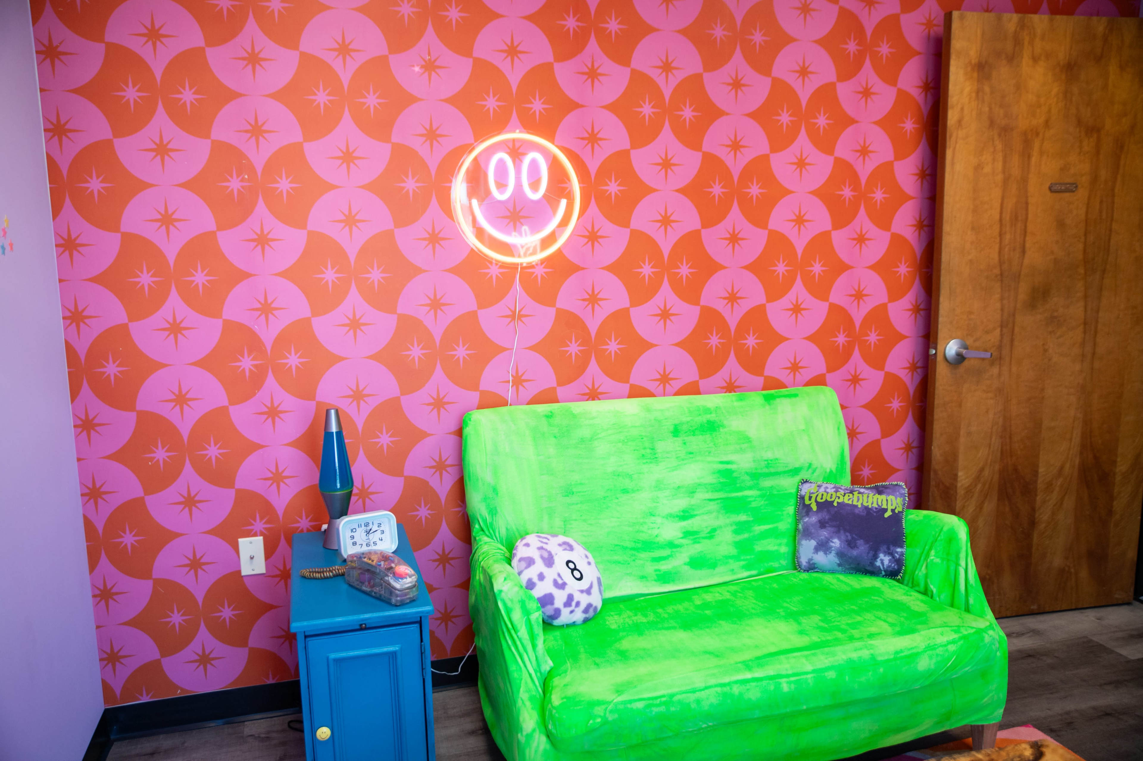 The image features a bright green couch placed against a pink and orange patterned wall, with a neon smiley face sign illuminated above.