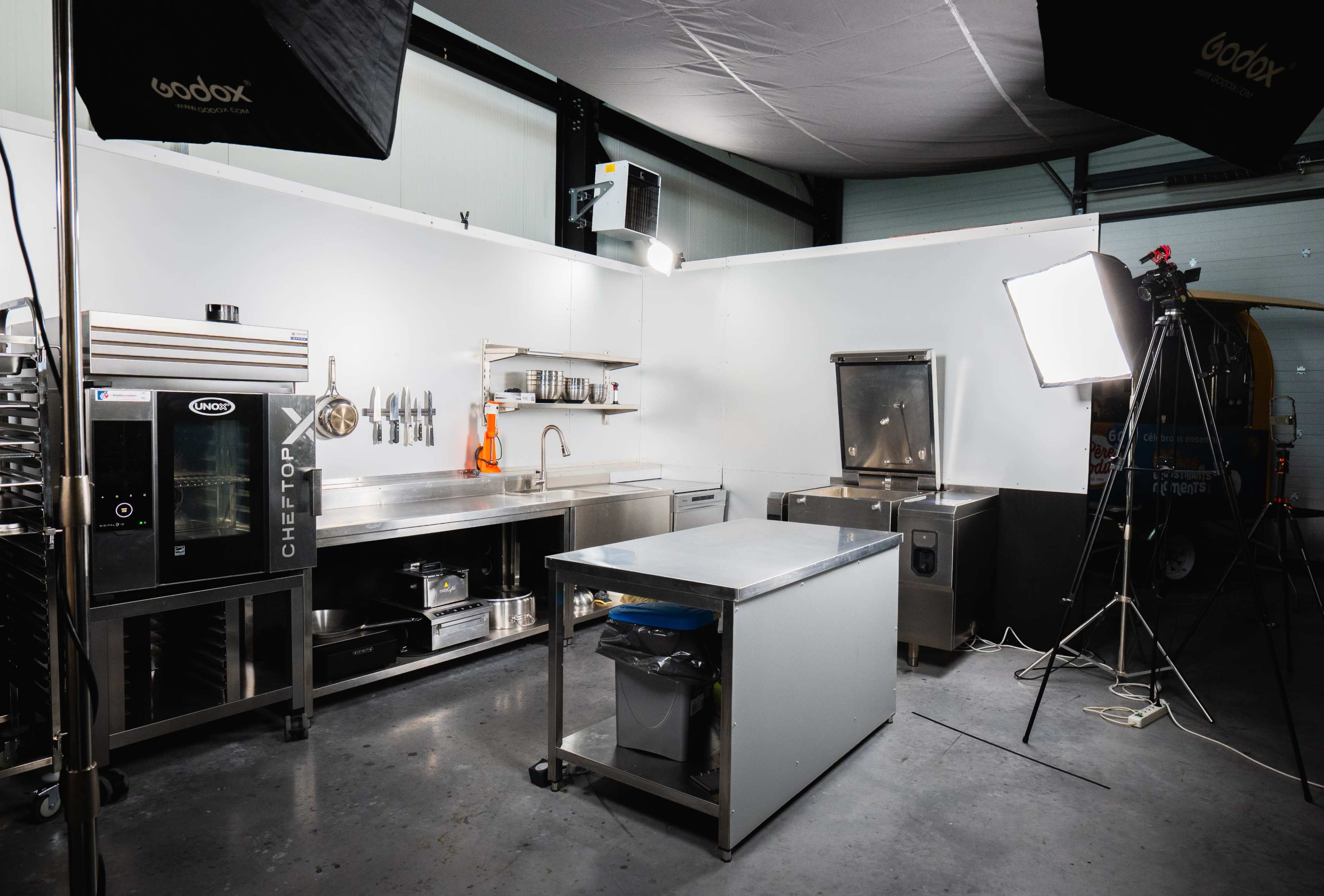 The image shows a commercial kitchen setting with stainless steel workstations, cooking equipment, and bright lighting.