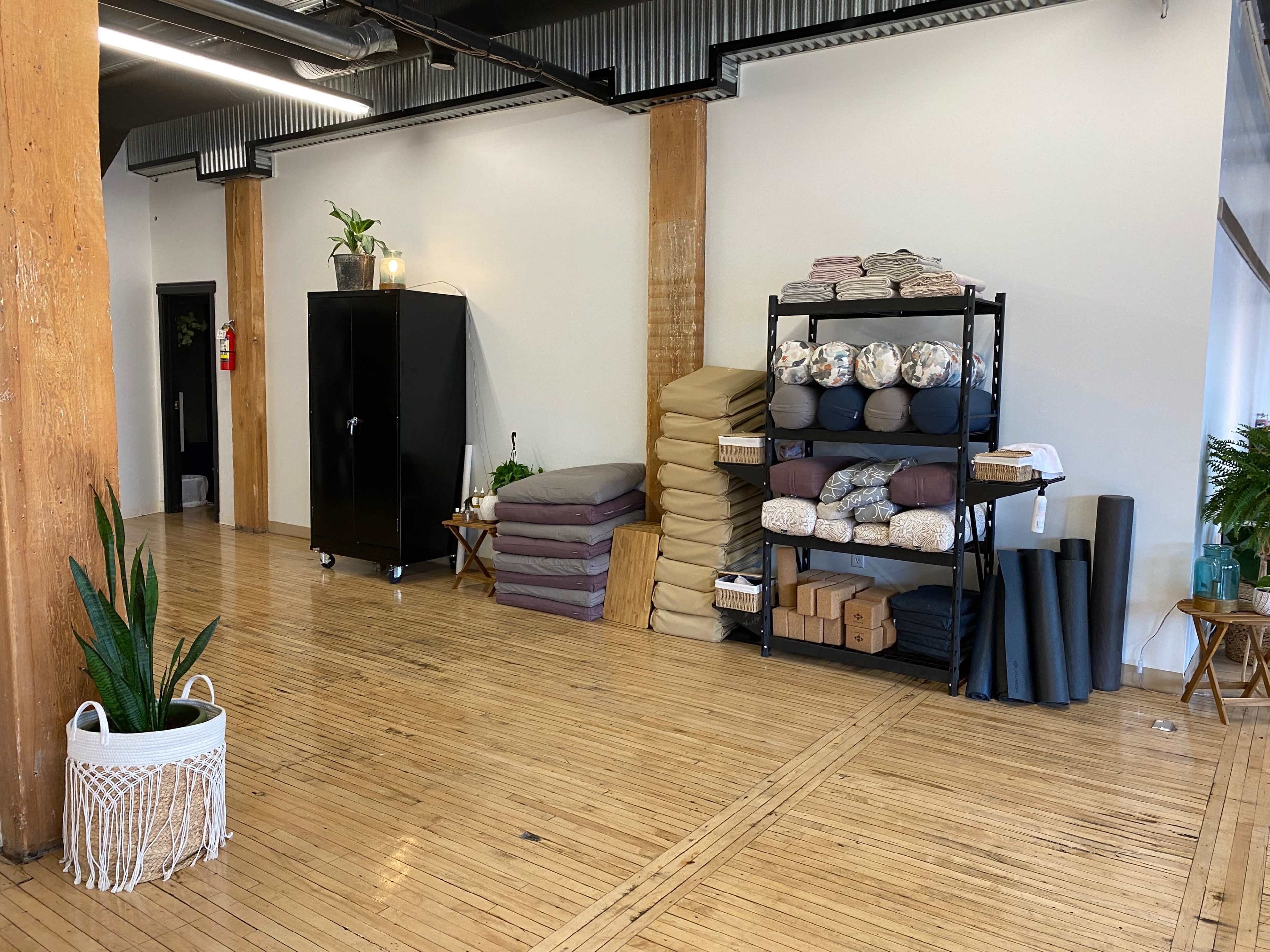 A spacious room with wooden flooring, featuring a black cabinet, stacked bedding materials, shelves with various items, and potted plants.