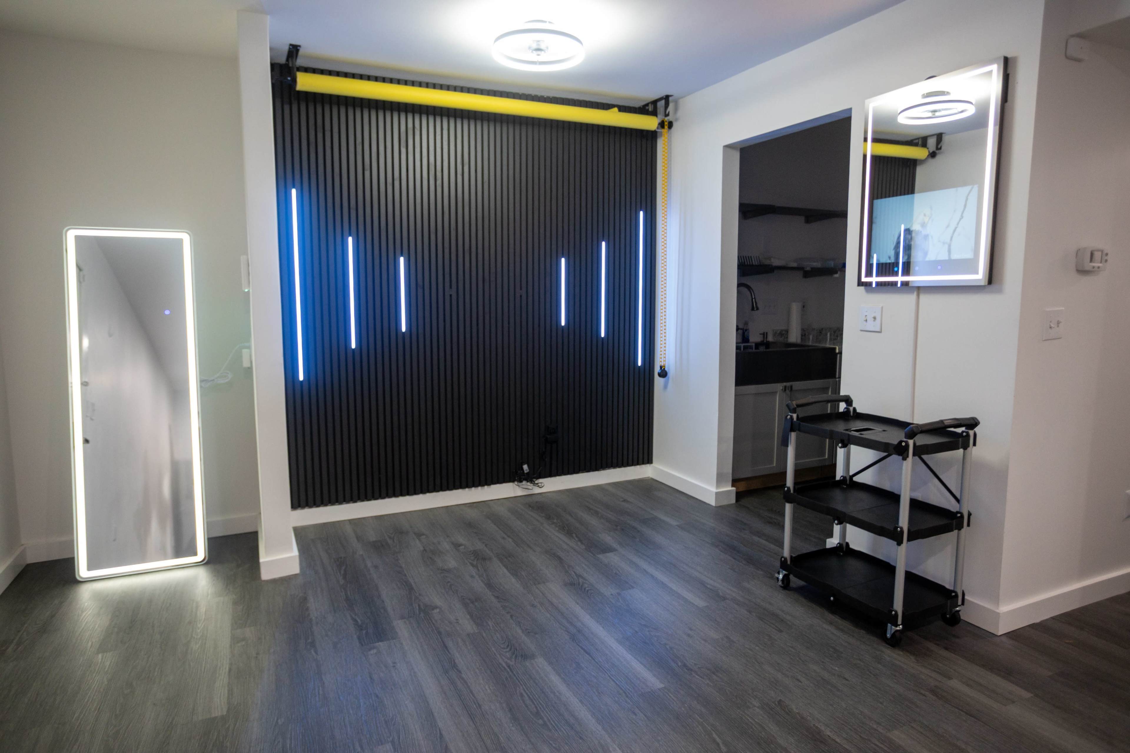 The image shows a modern room featuring a black wall with vertical LED light strips, a sleek full-length mirror, and a small rolling cart.