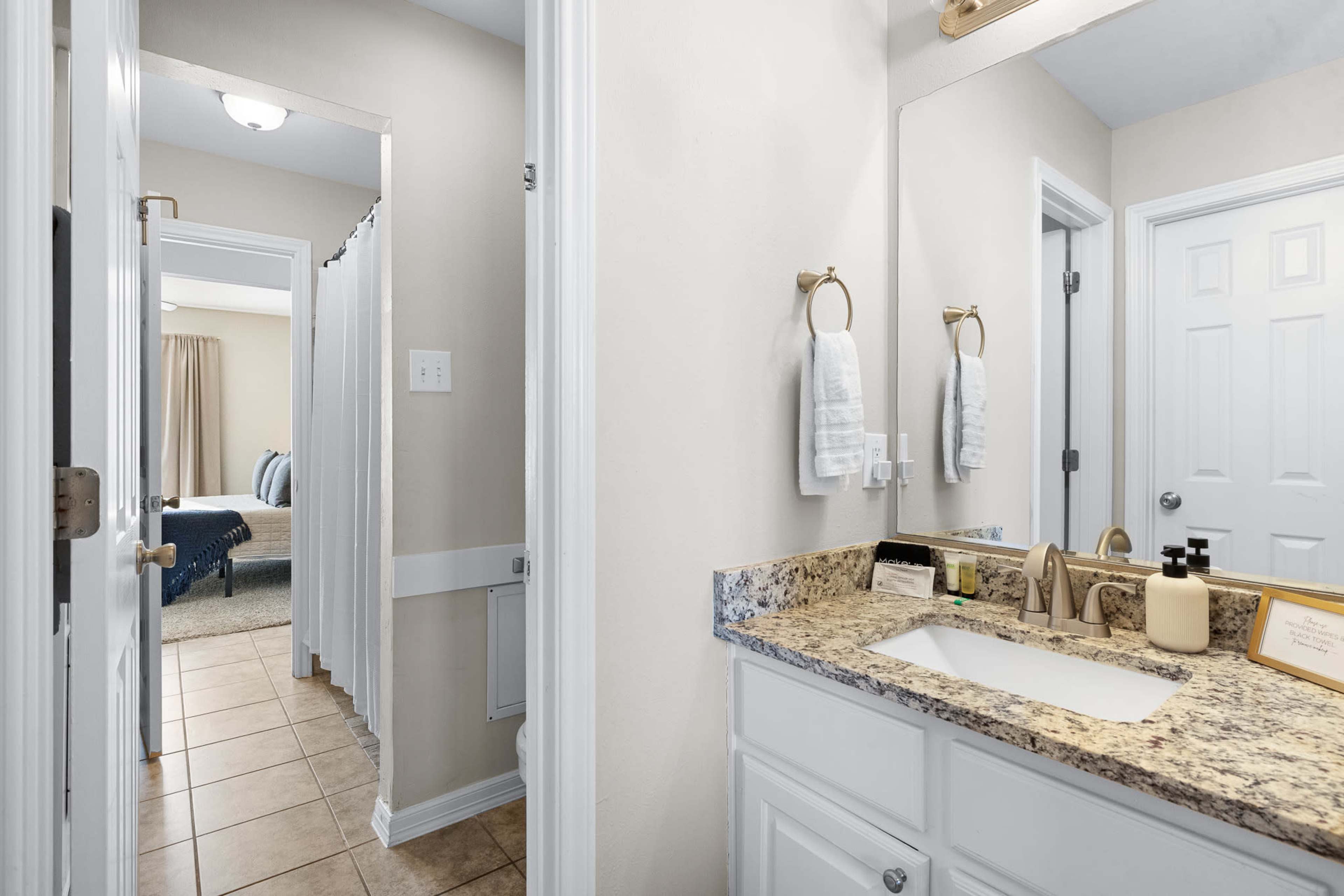 The image shows a bathroom with a granite countertop, dual sinks, and a door leading to a hallway and a bedroom.