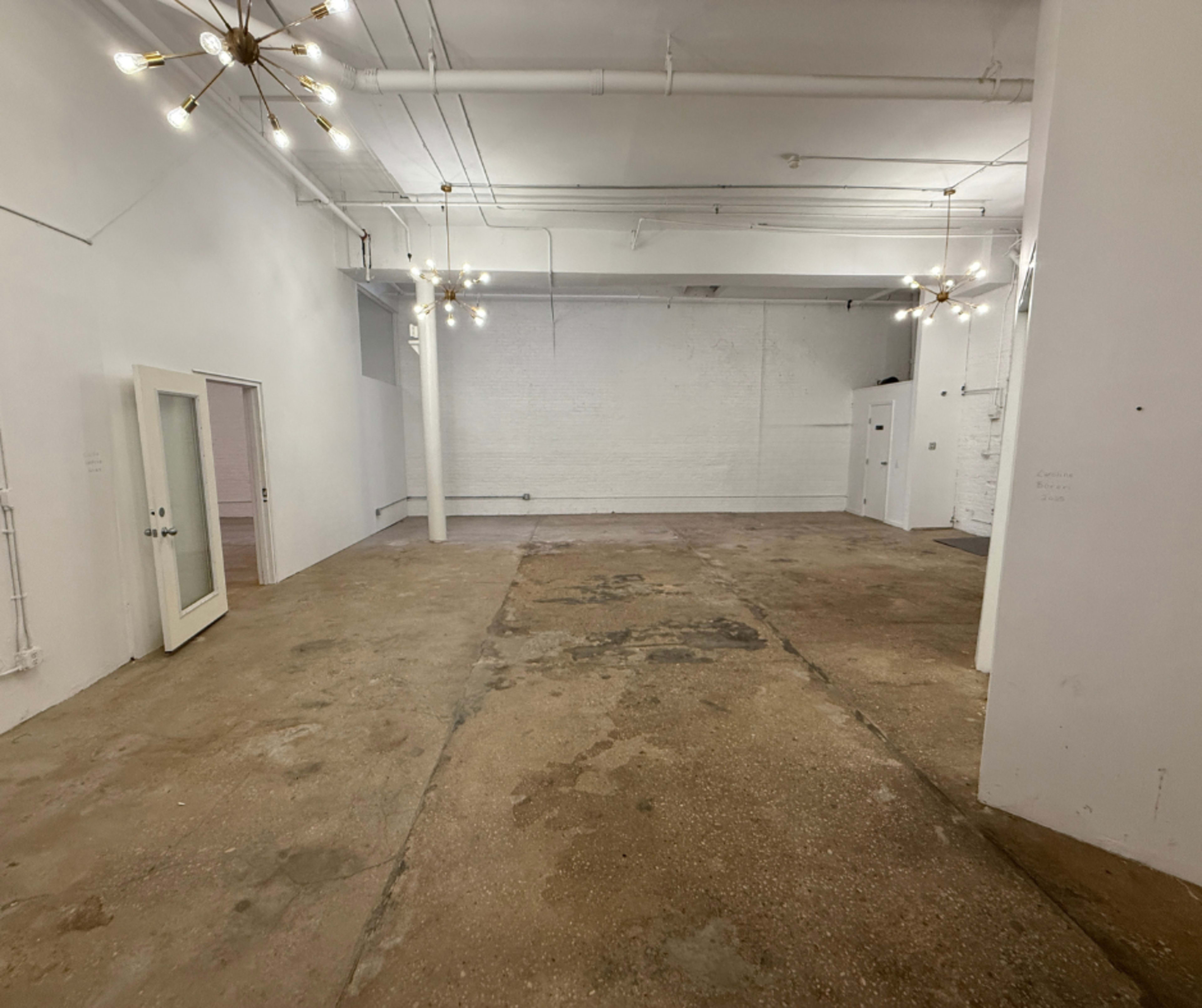 The image shows a spacious, empty room with a concrete floor, exposed pipes, and minimalist lighting fixtures.