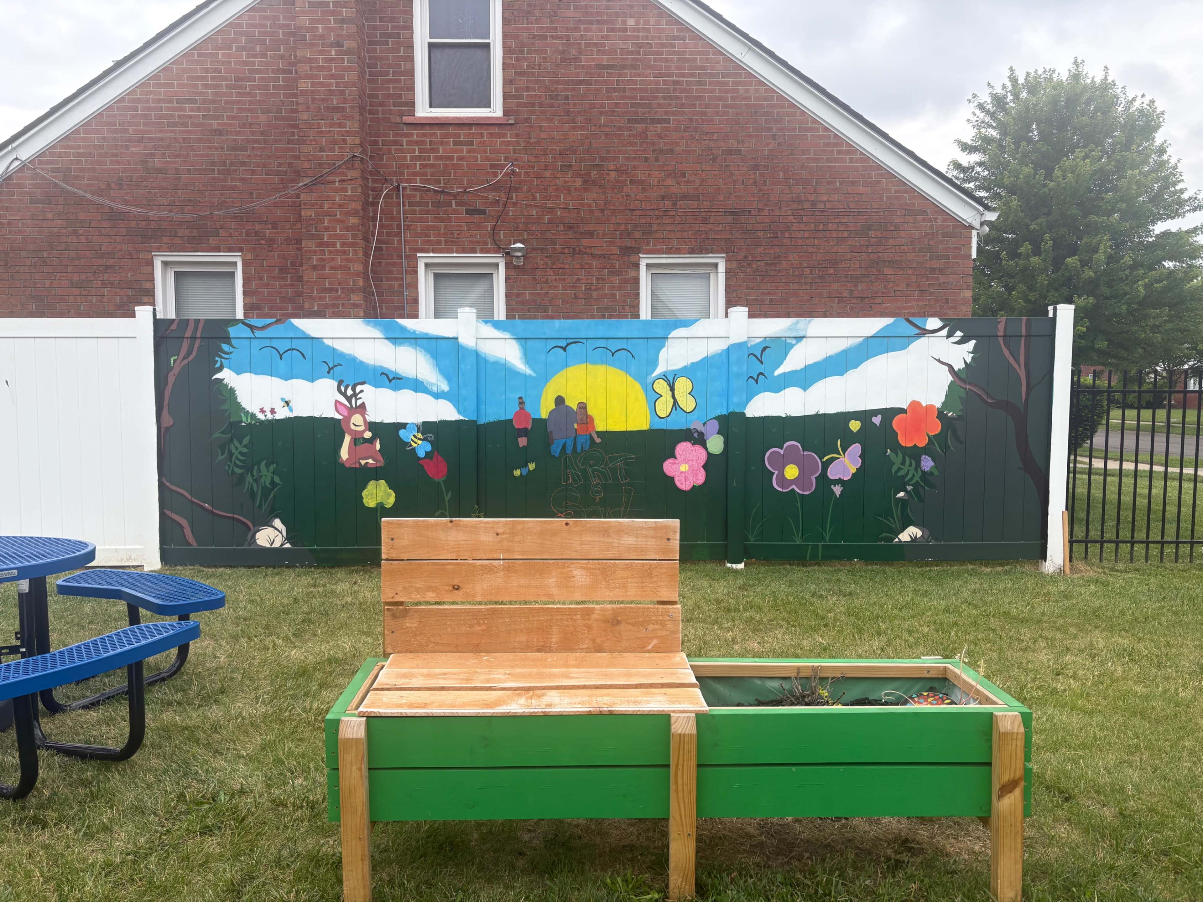 A wooden bench is positioned in front of a colorful mural depicting a landscape with clouds, a sun, and flowers on a fence near a brick building.