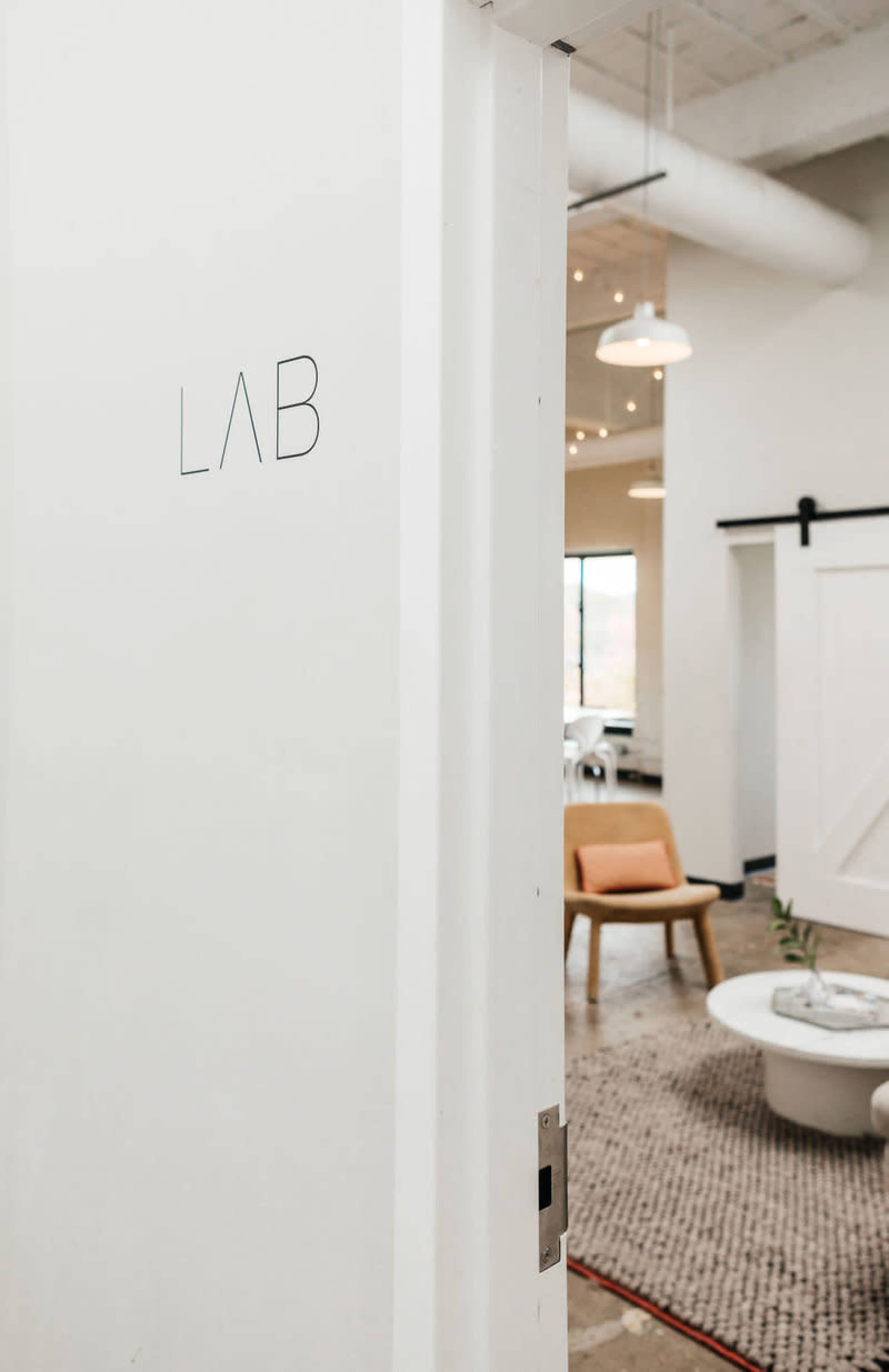 The image shows an open door labeled "LAB" leading into a modern, well-lit space with minimalist furniture.