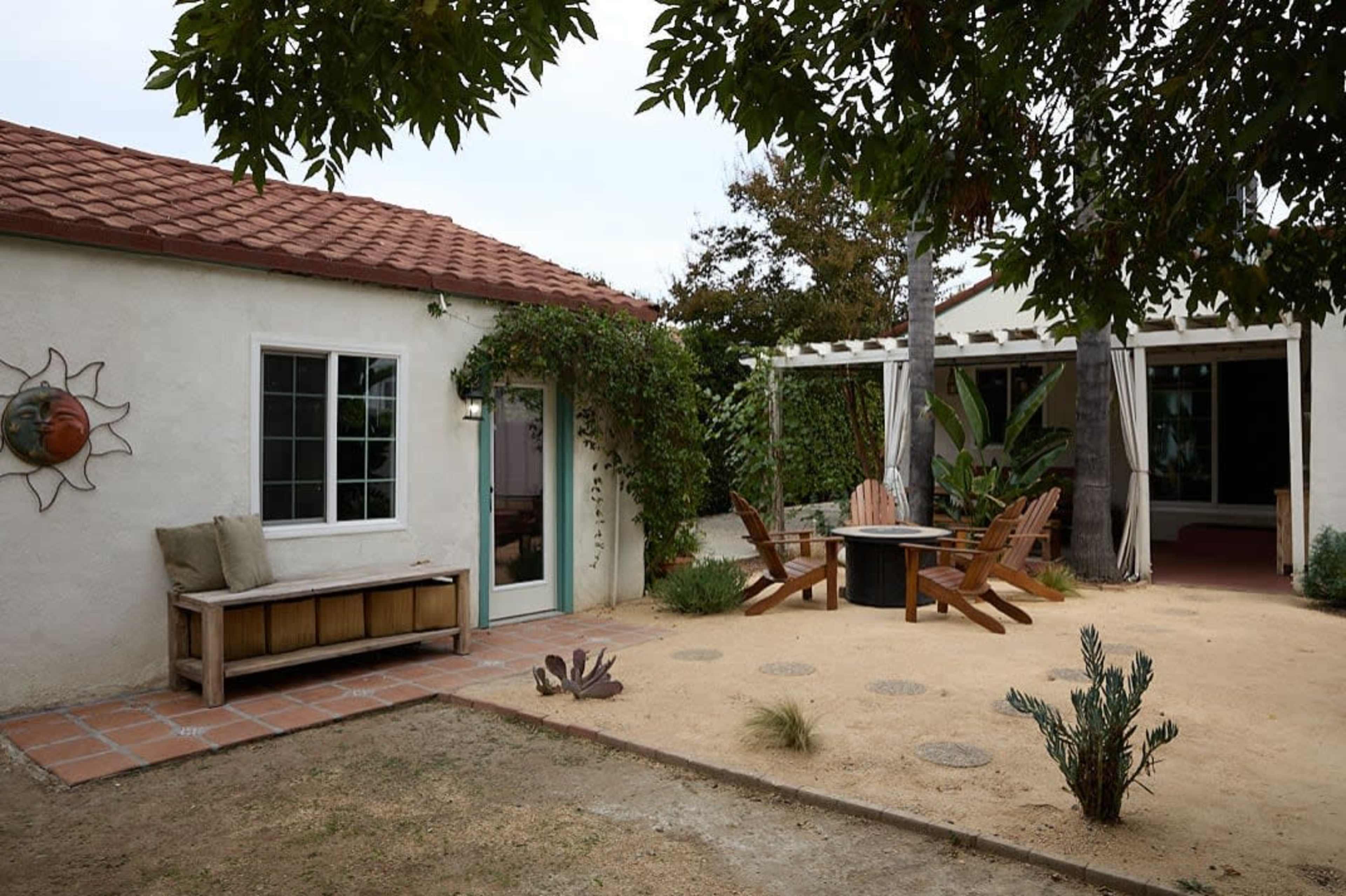The image shows a cozy outdoor space featuring a patio with wooden chairs, a small table, and surrounding greenery near a house with a red-tiled roof.