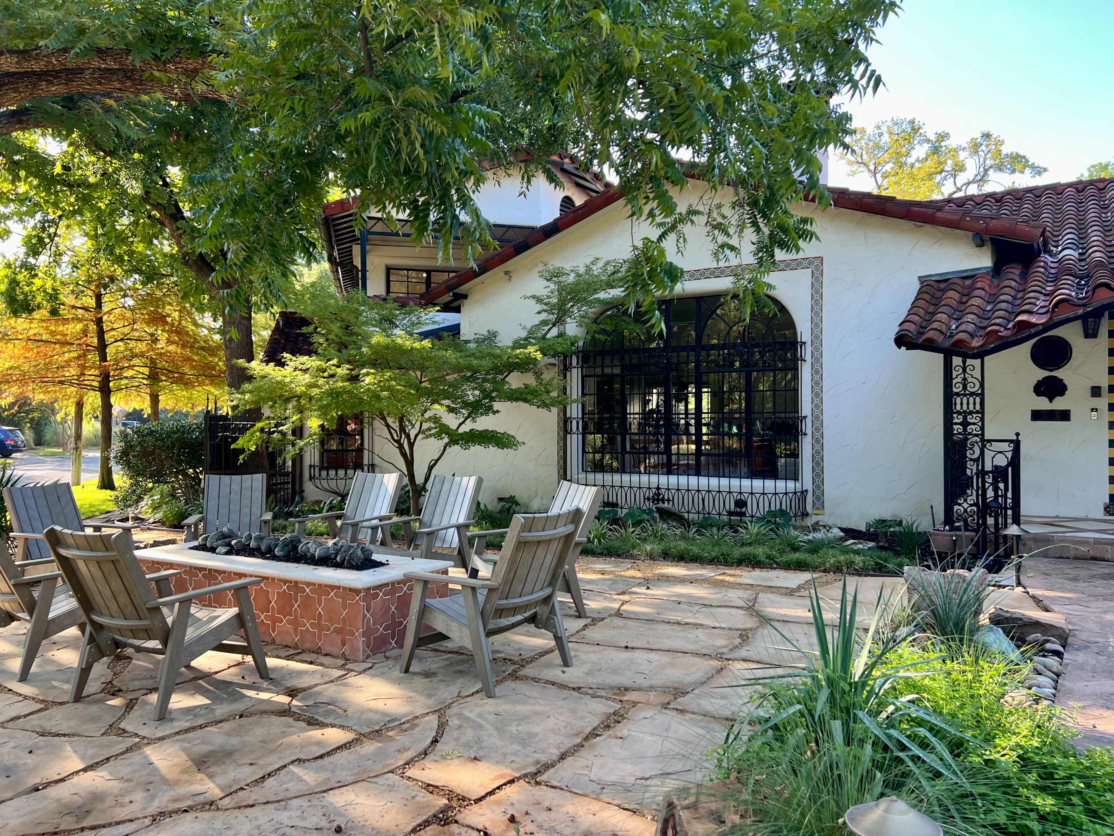 A charming patio area with wooden chairs arranged around a fire pit in front of a white stucco house surrounded by greenery.