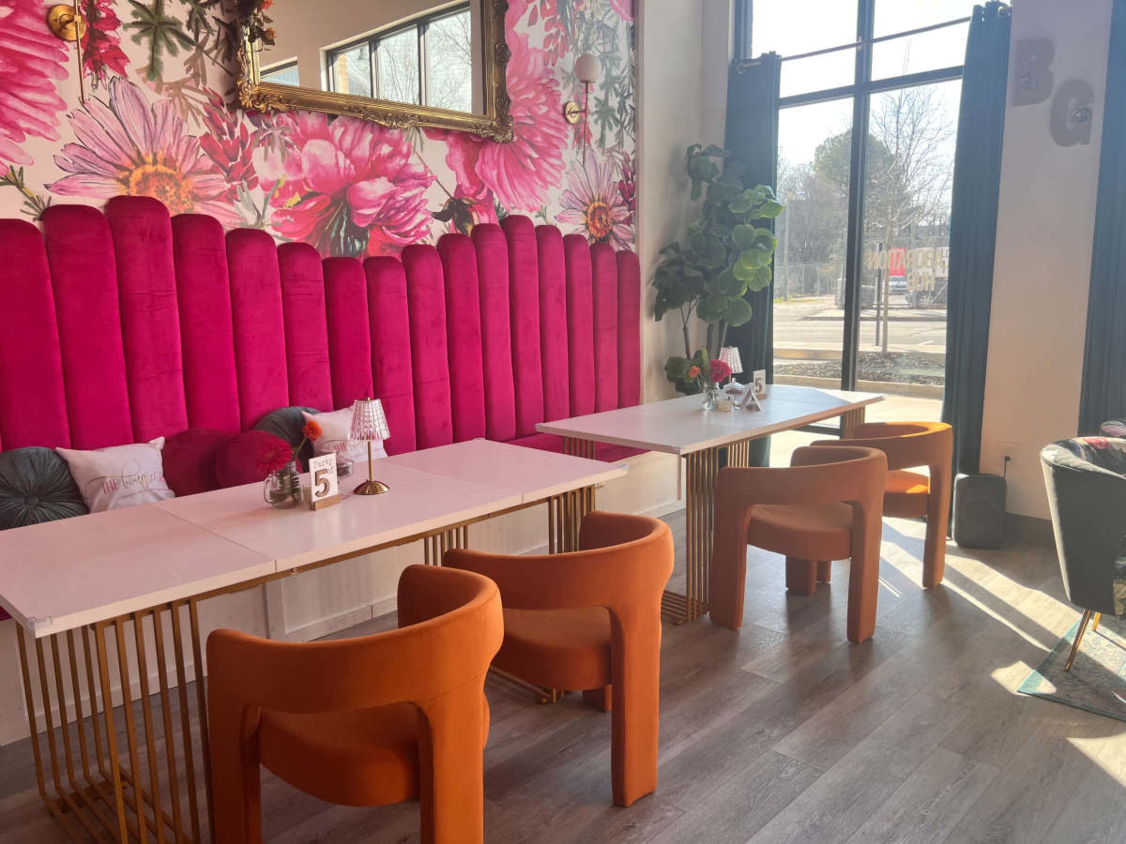 The image shows a brightly decorated café with pink floral wallpaper, two tables with white tops and gold legs, and orange velvet chairs.