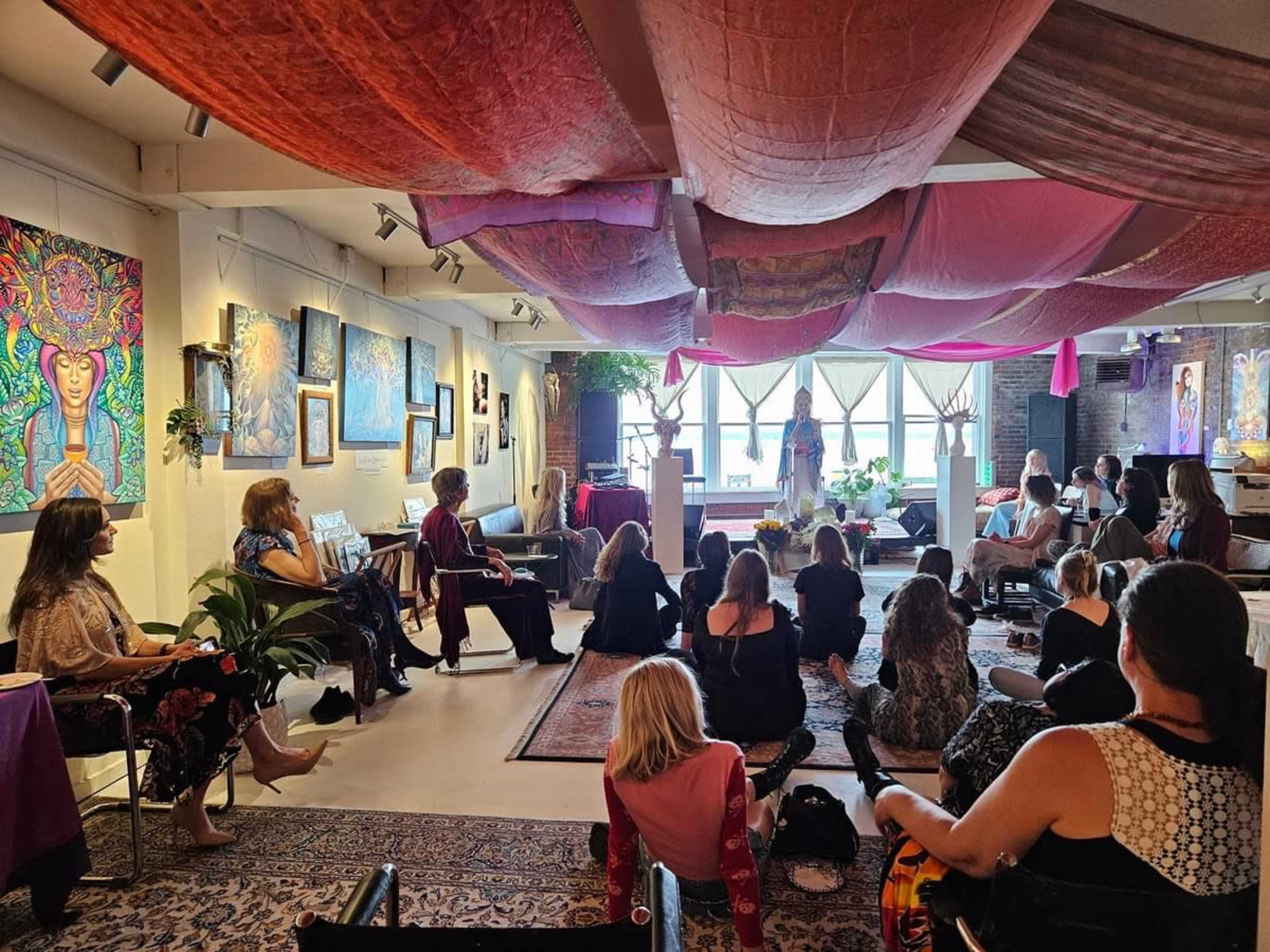 A diverse group of people sit on the floor and in chairs, listening to a speaker in a colorful, art-filled room with fabric draped from the ceiling.