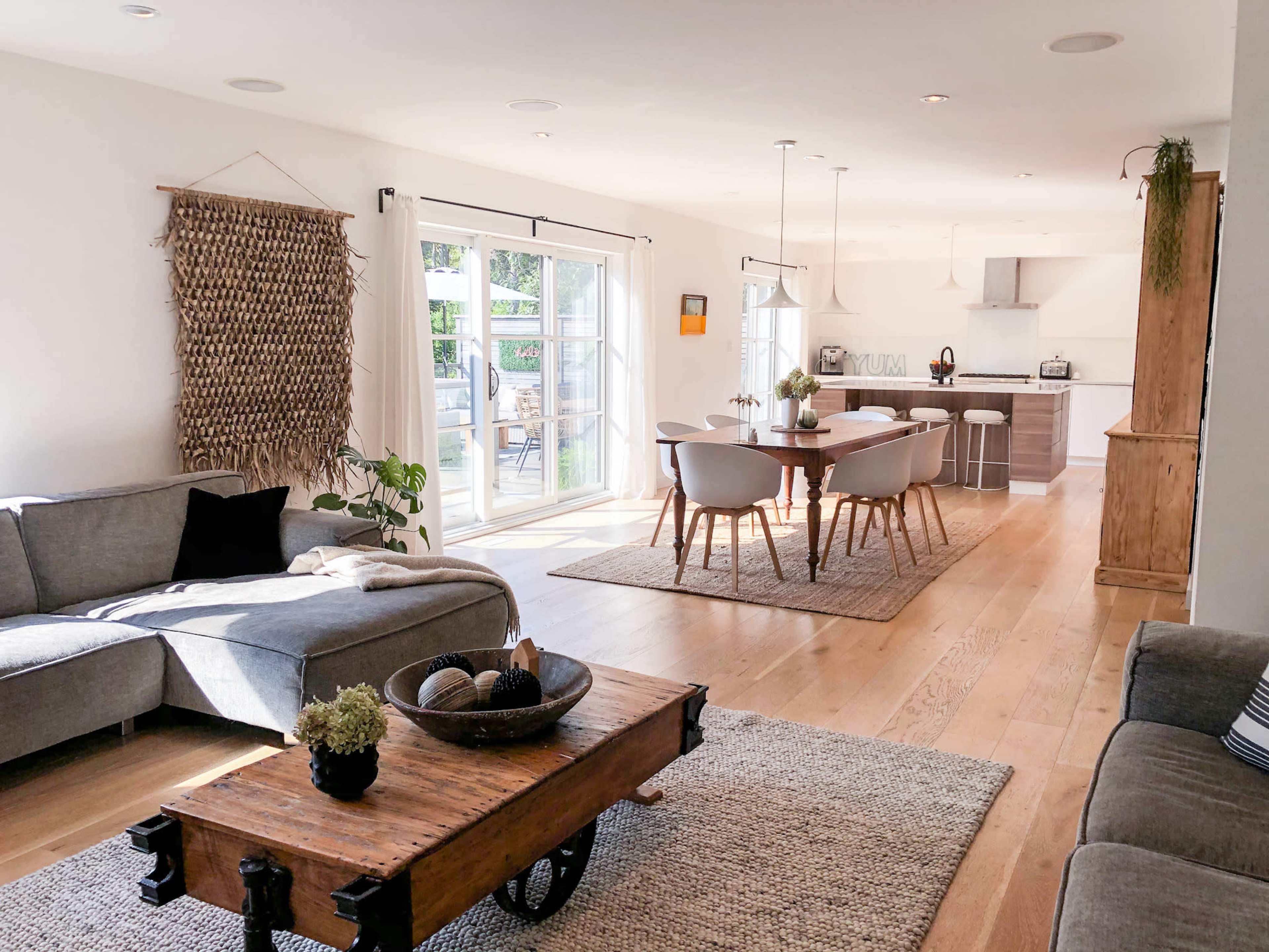 A bright, open living space featuring a gray sofa, a wooden coffee table on a textured rug, and a dining area with light wood chairs and a modern kitchen in the background.