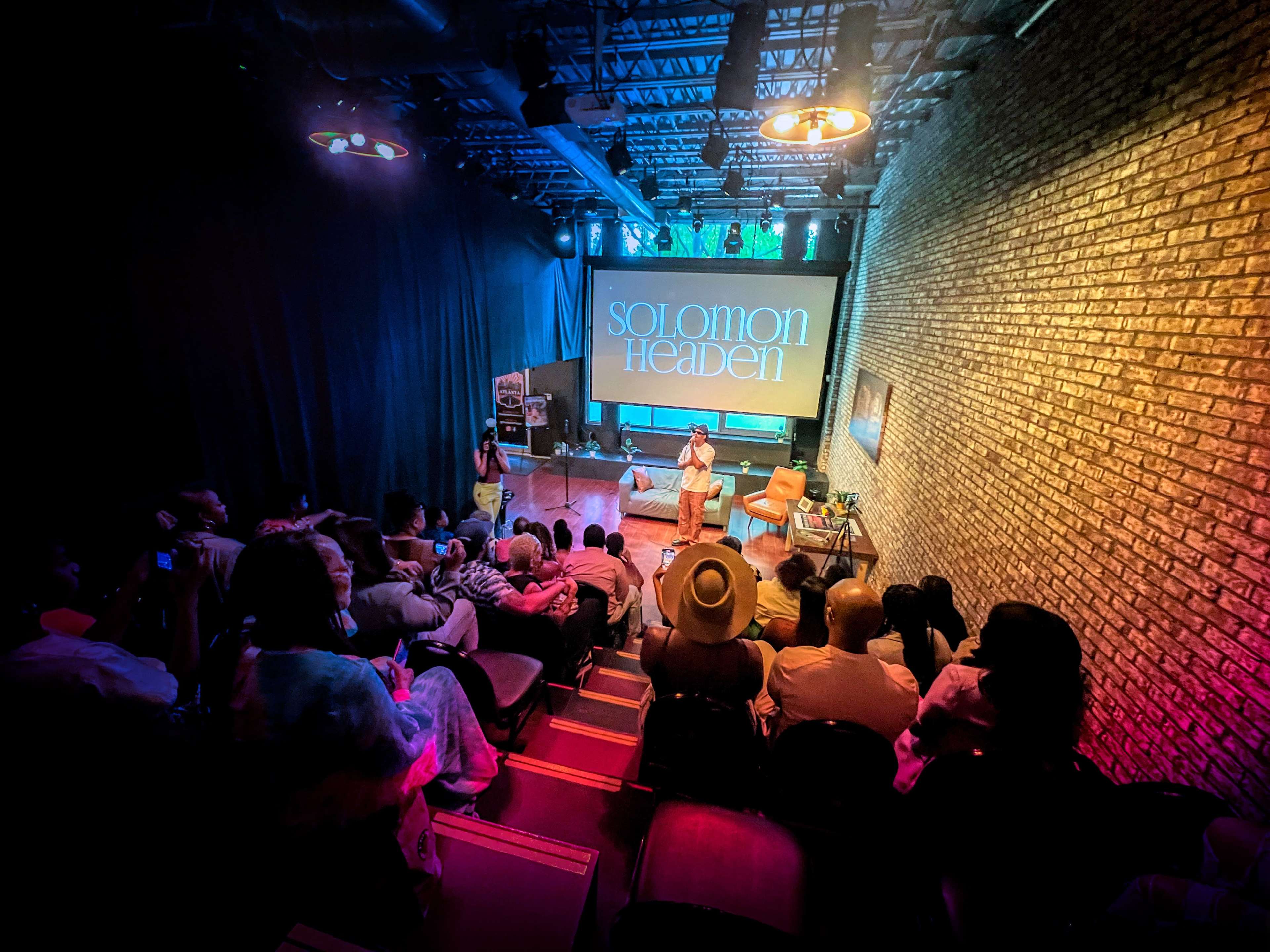 A small audience gathers in a dimly lit theater as a speaker addresses the crowd, with a large screen displaying "SOLOMON HEADEN" in the background.