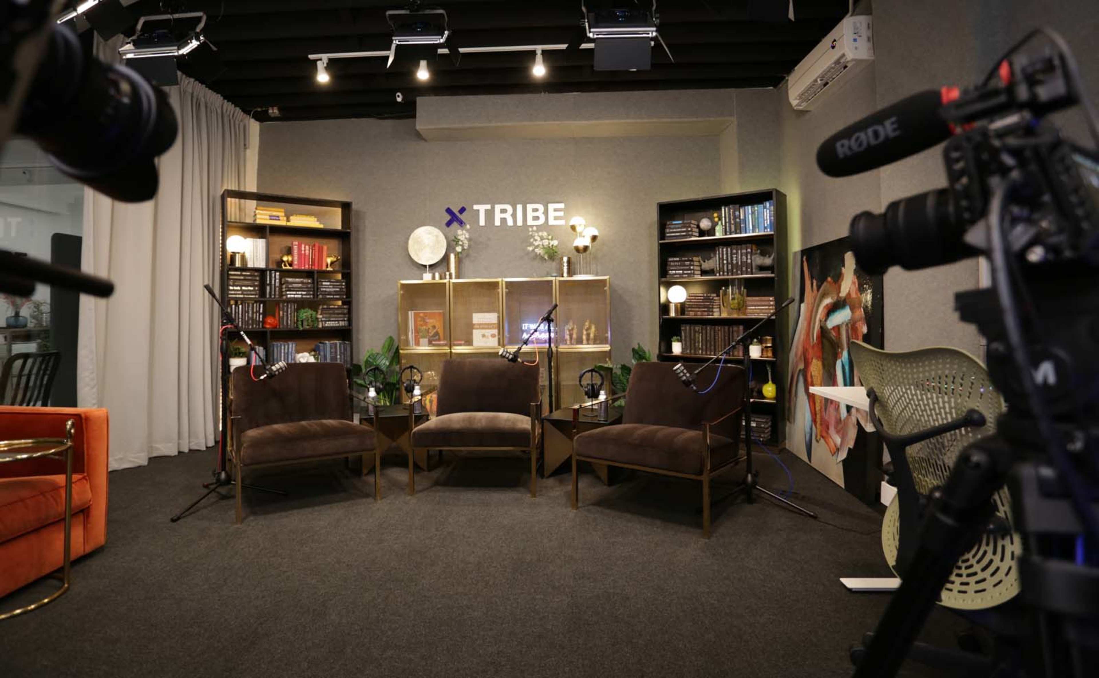 A modern interview setup with three brown chairs facing a decorative backdrop featuring shelves with books and plants, alongside microphones and cameras in the foreground.
