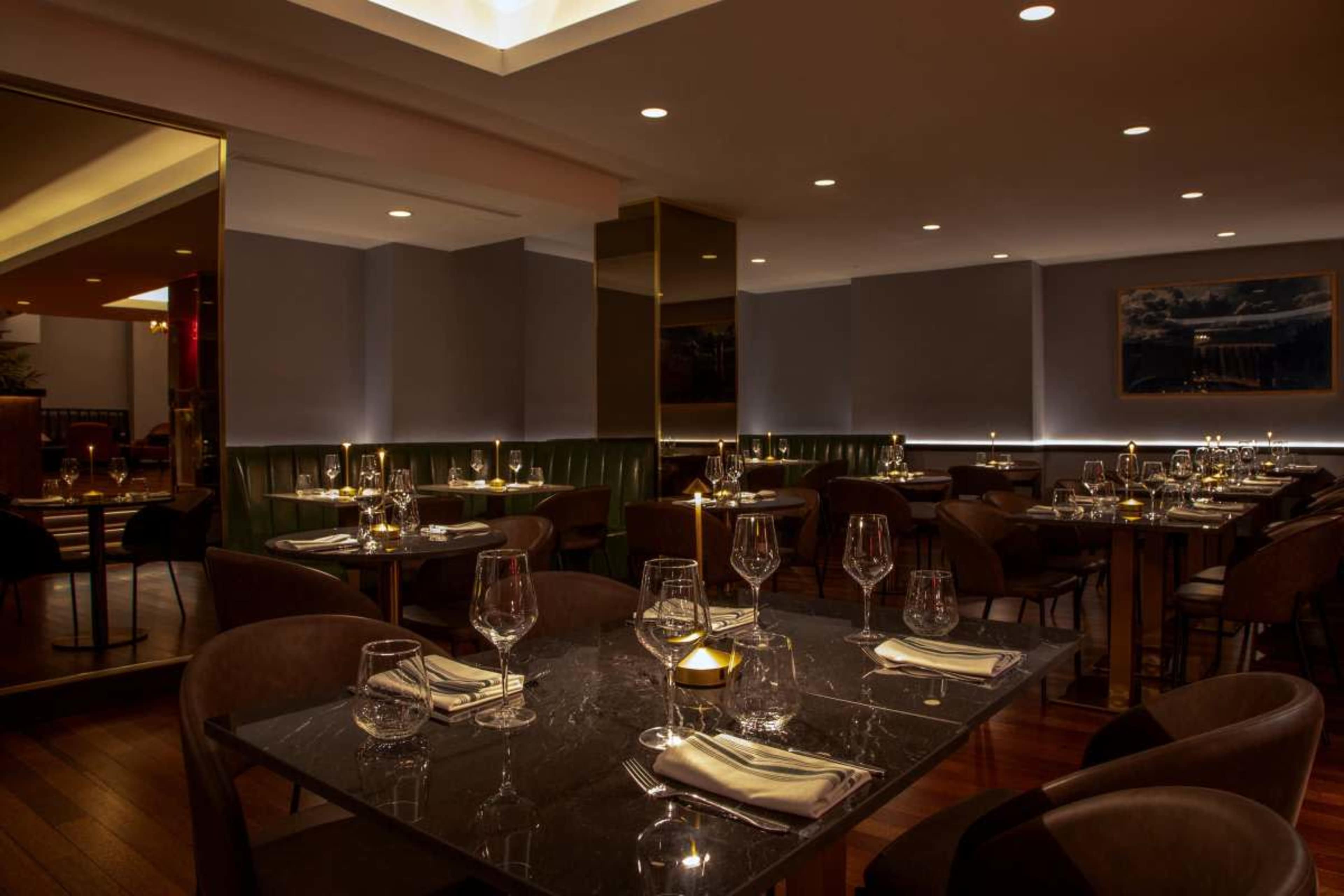 The image shows a modern restaurant interior with elegantly set tables, soft lighting, and a mix of dark and neutral colors.