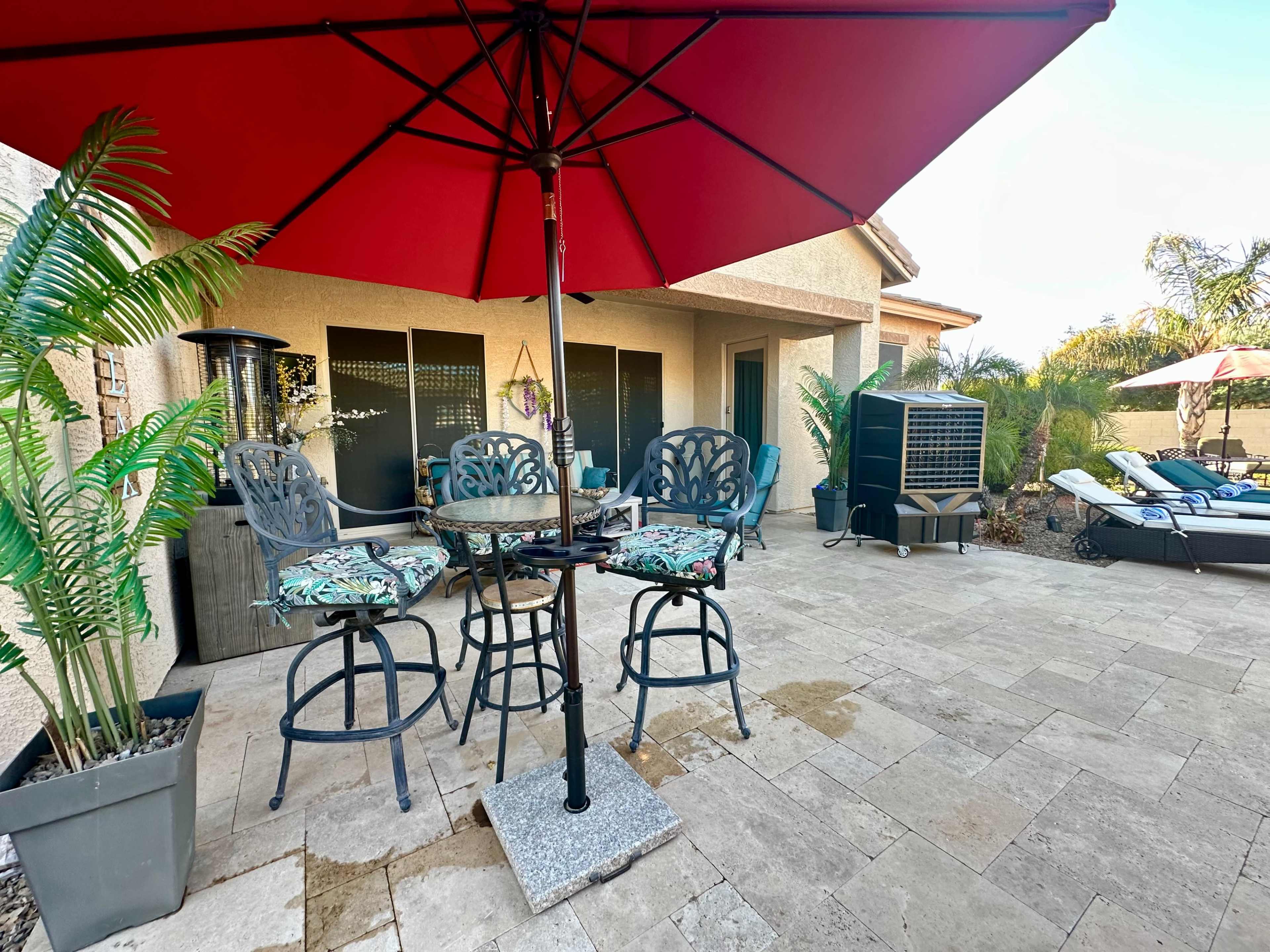 A patio area features a round table with high stools under a red umbrella, surrounded by potted plants and lounge chairs.