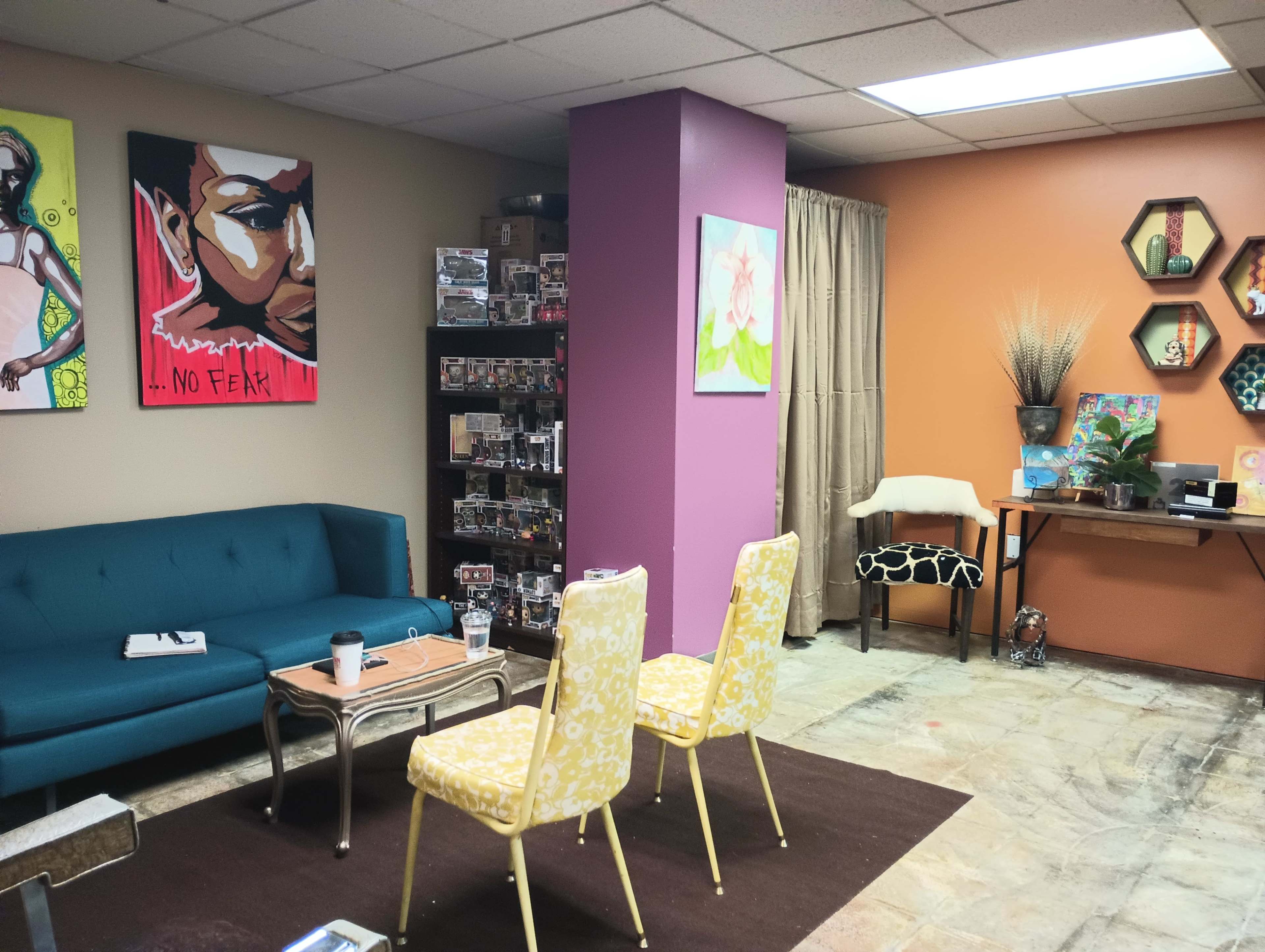 The image shows a cozy room with a teal sofa, two yellow chairs, colorful artwork on the walls, and various decorative items on shelves and tables.