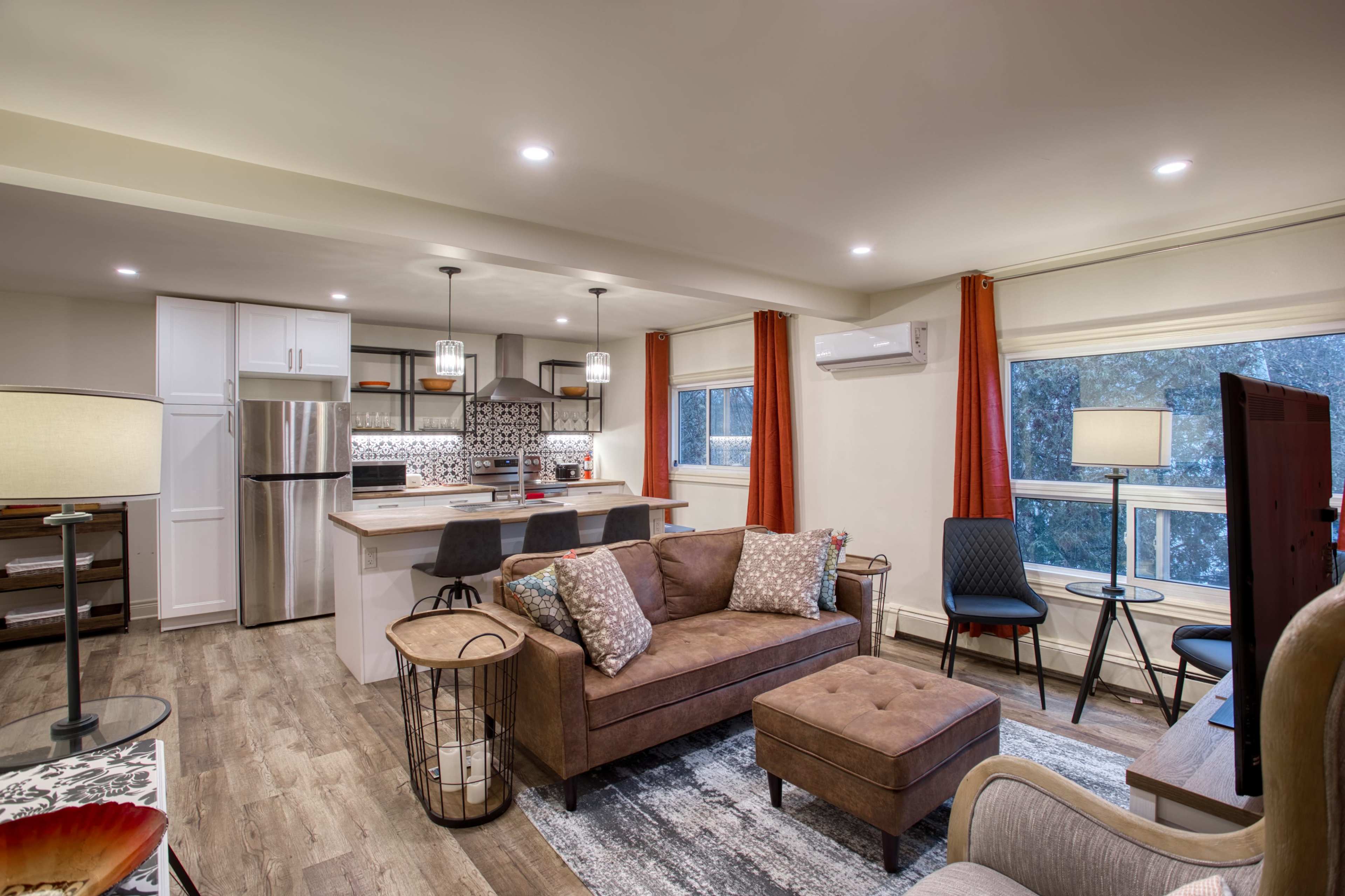 The image shows a modern living room and kitchen area featuring a brown sofa, a small ottoman, kitchen bar with stools, and large windows with orange curtains.