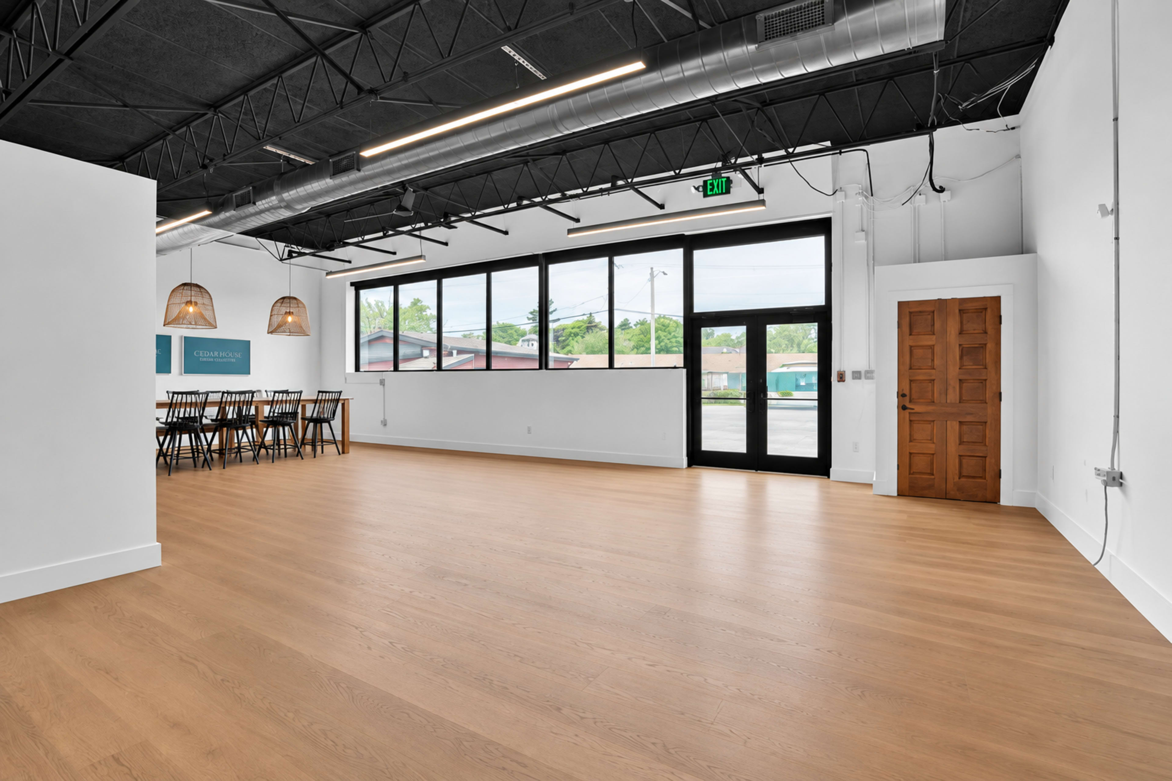 The image shows an empty, modern interior space with wooden flooring, large windows, and a dining area with a long table and chairs.