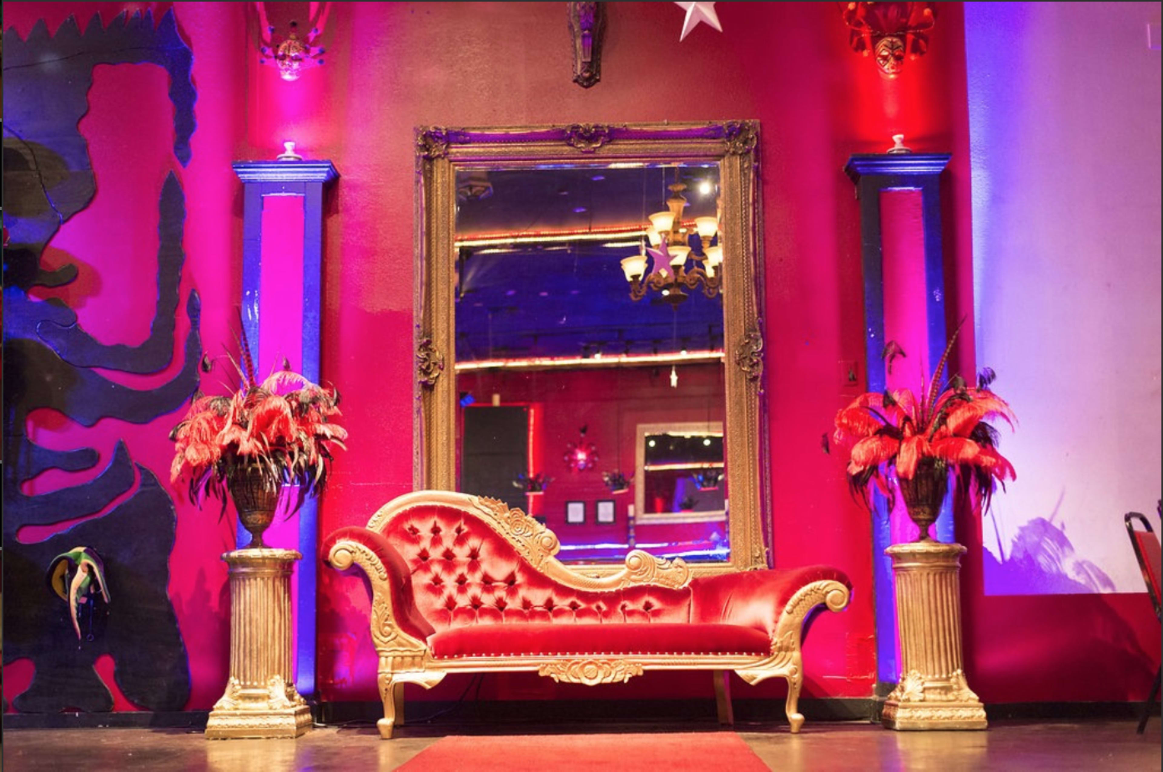The scene features a red and gold decorative sofa positioned in front of a large ornate mirror, with vibrant red and purple lighting illuminating the walls and two tall floral arrangements on either side.