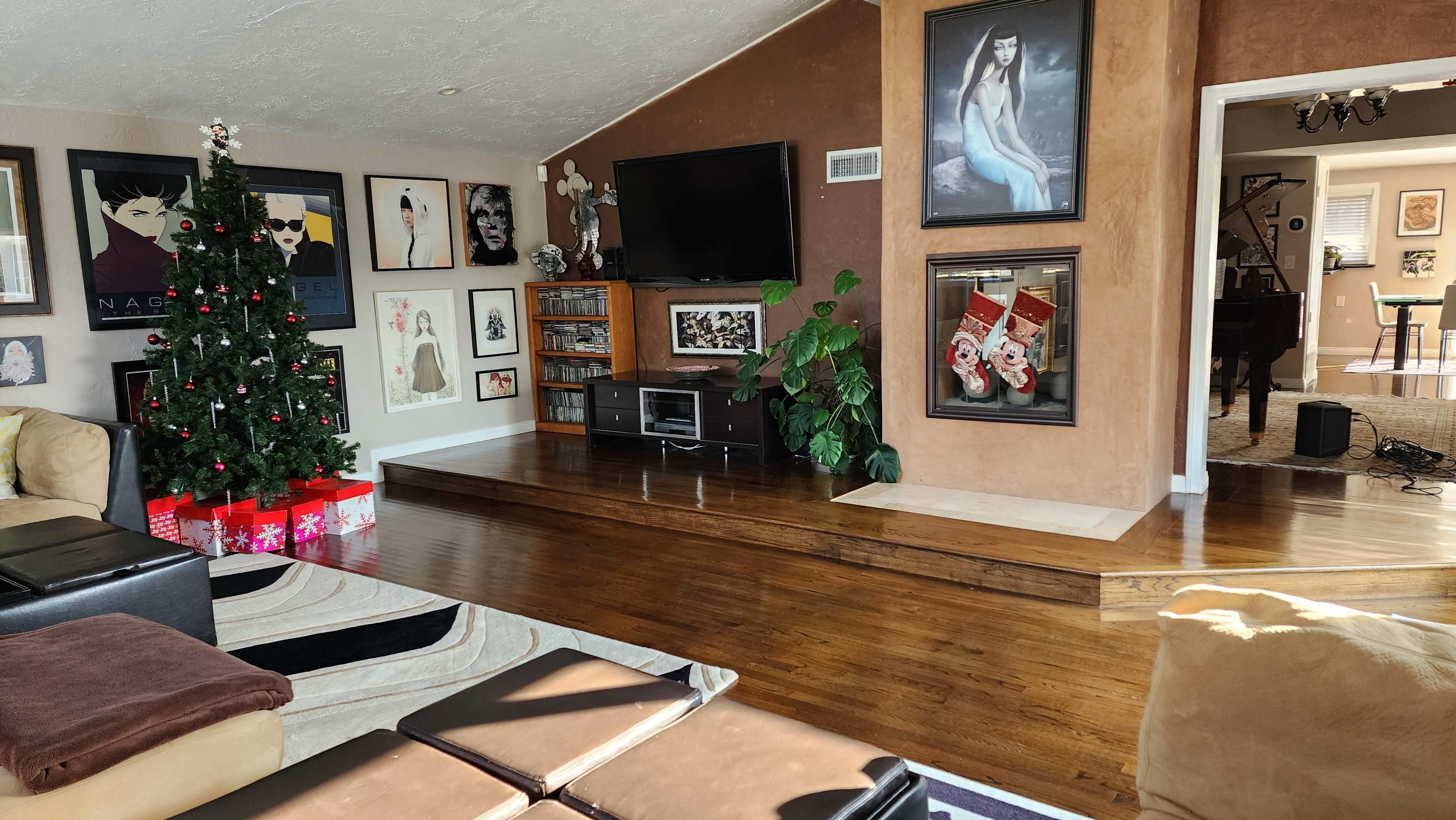 The image shows a living room featuring a Christmas tree, a television mounted on the wall, and a variety of framed artworks arranged around the space.