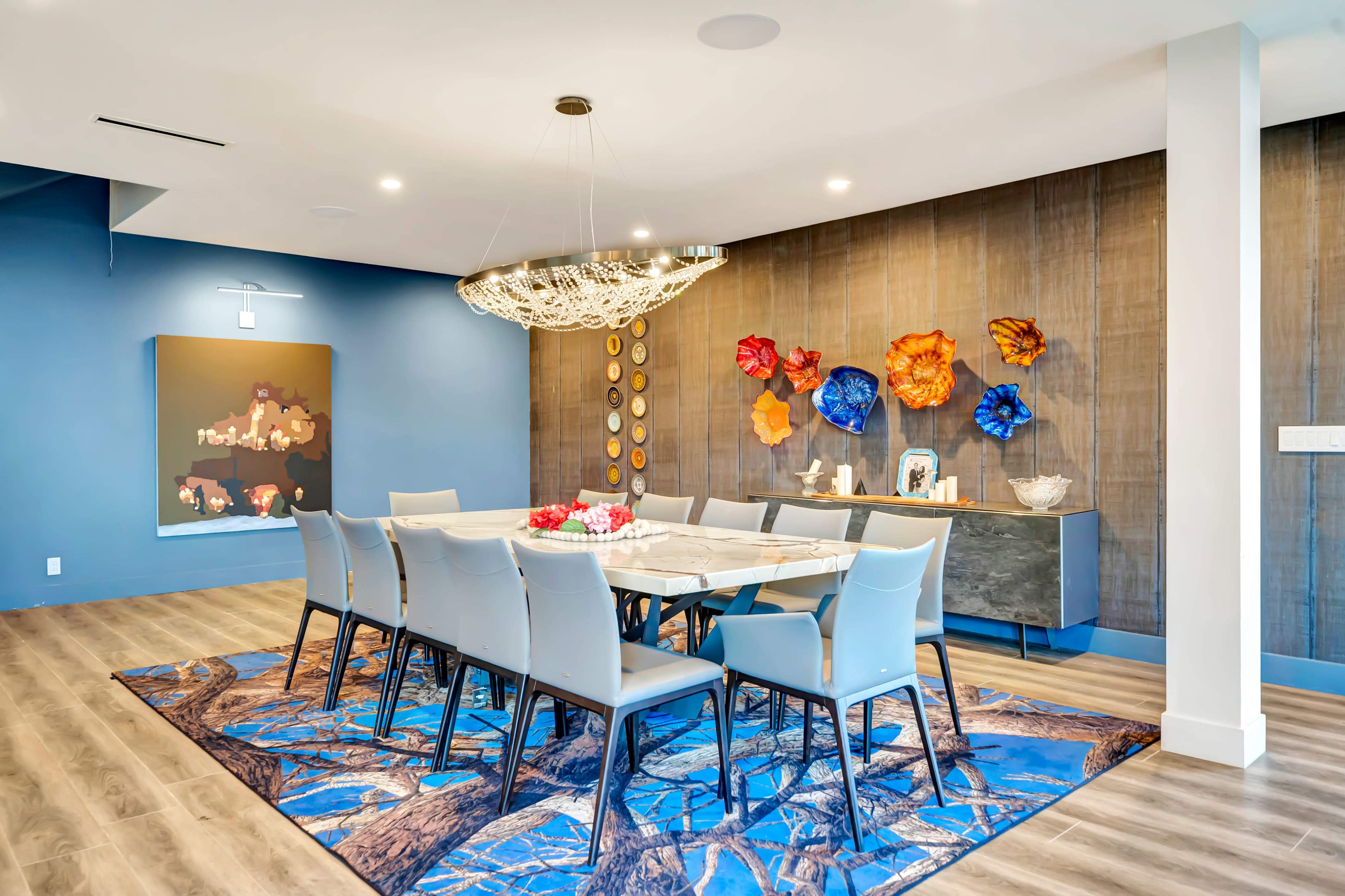 A dining room features a long table surrounded by ten chairs, with decorative wall art and a large chandelier above.