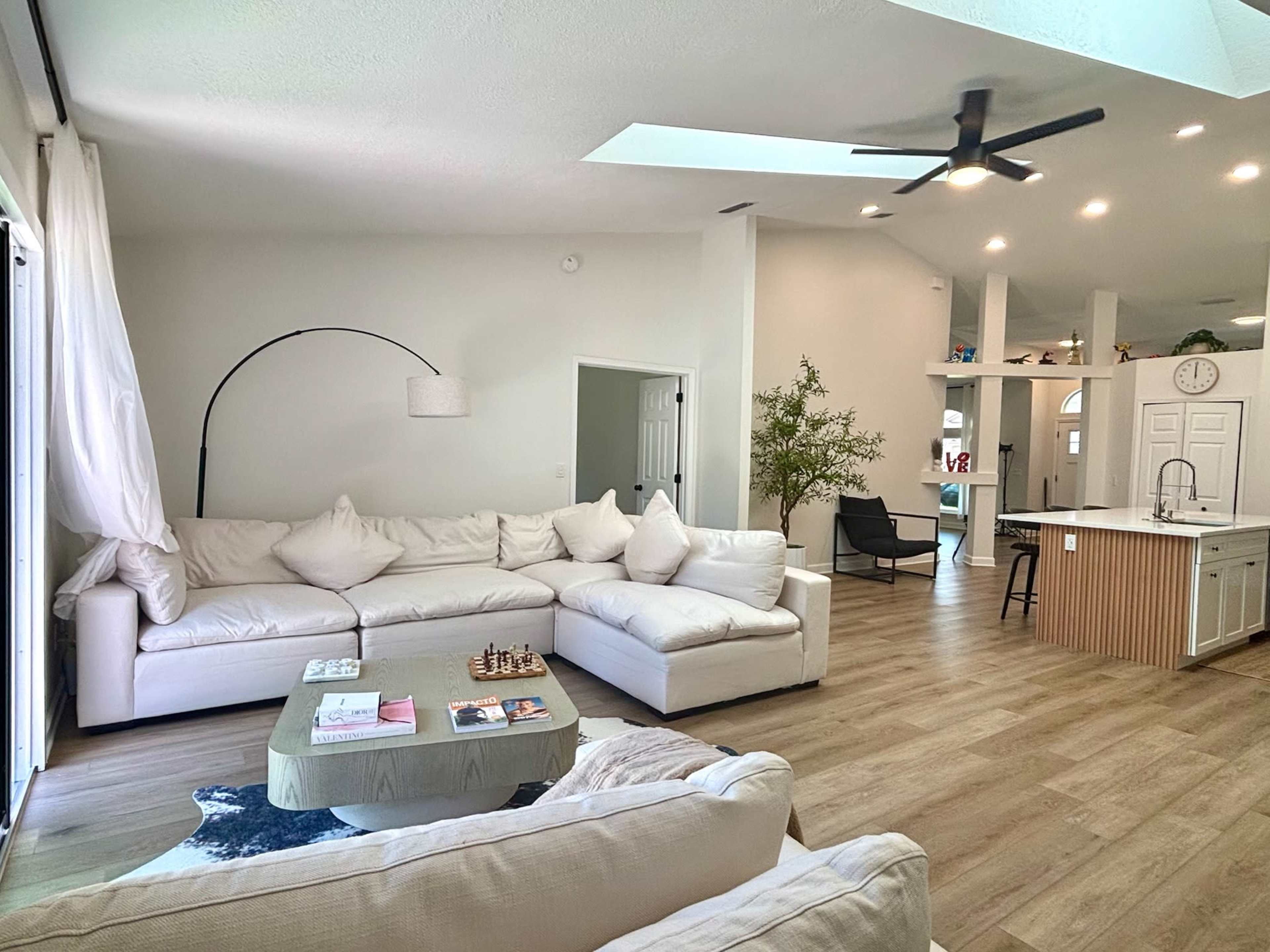 The image shows a spacious, modern living room with a large white sectional sofa, a green coffee table, and an open kitchen area in the background.