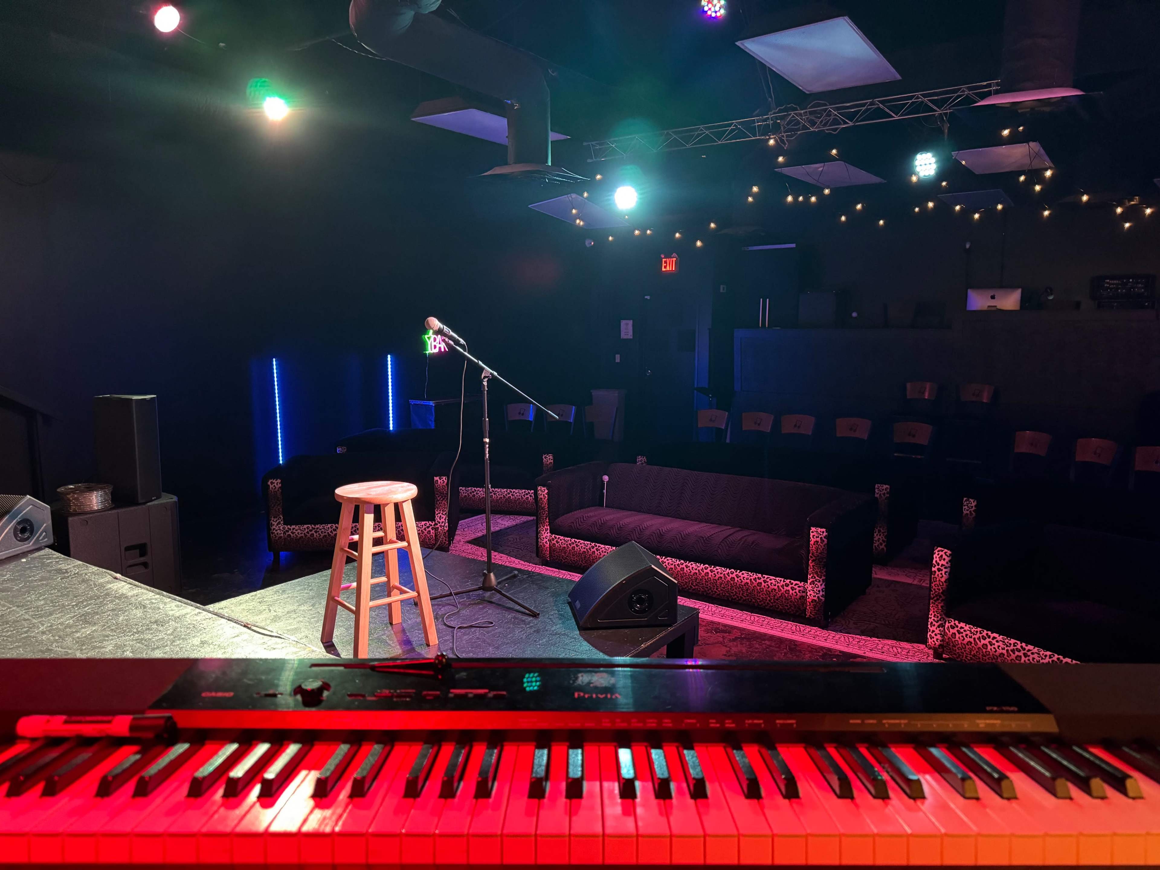 A stage view of a small performance venue with a microphone on a stand, a wooden stool, and a keyboard in the foreground, surrounded by couches and stage lights.
