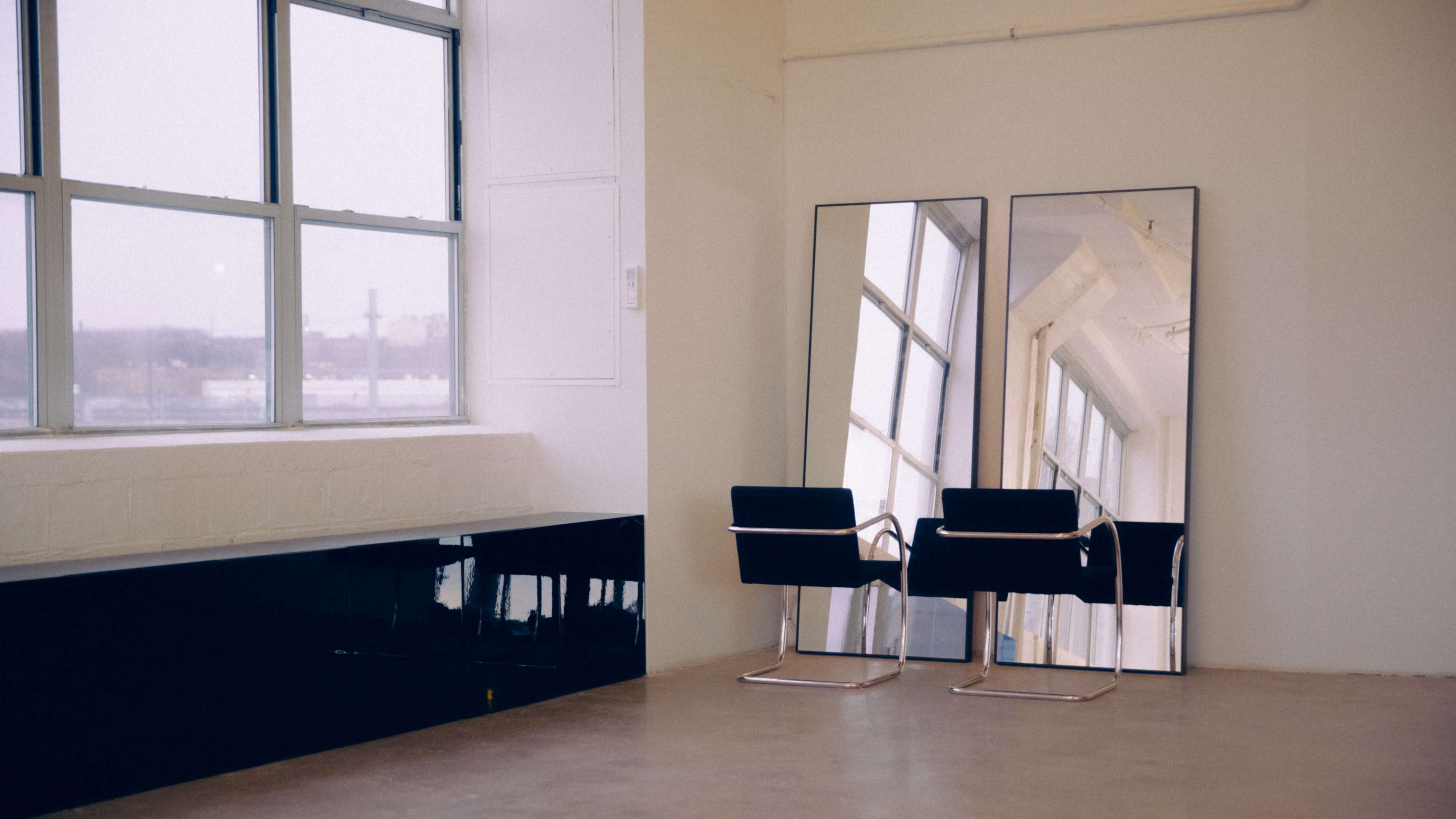 The image shows a minimalist interior space with two black chairs positioned next to large, framed mirrors and a window revealing a hazy view outside.